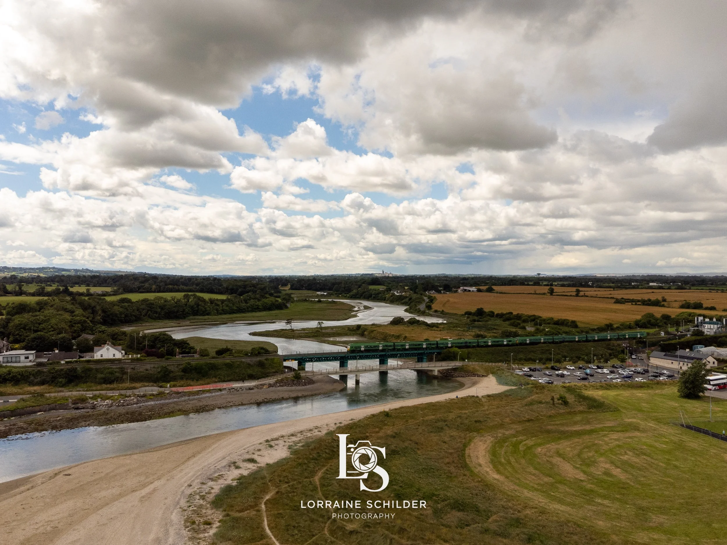 A scenic landscape showing a river with a bridge, a train passing over it, surrounded by green fields, a parking lot, houses, and a partly cloudy sky. Laytown, Meath.