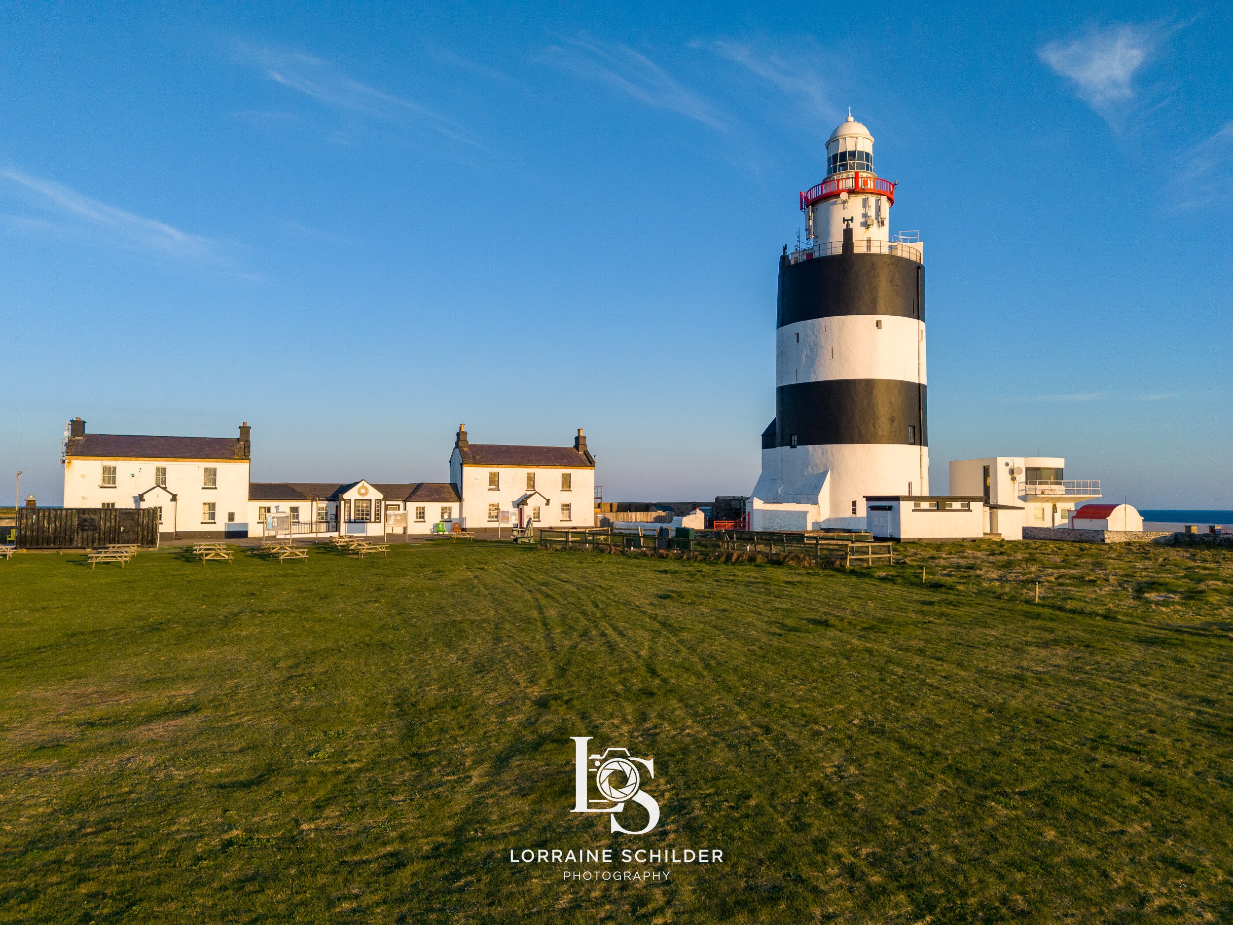 A black, white, and red lighthouse standing tall near a few white buildings on a grassy area under a blue sky. Wexford.