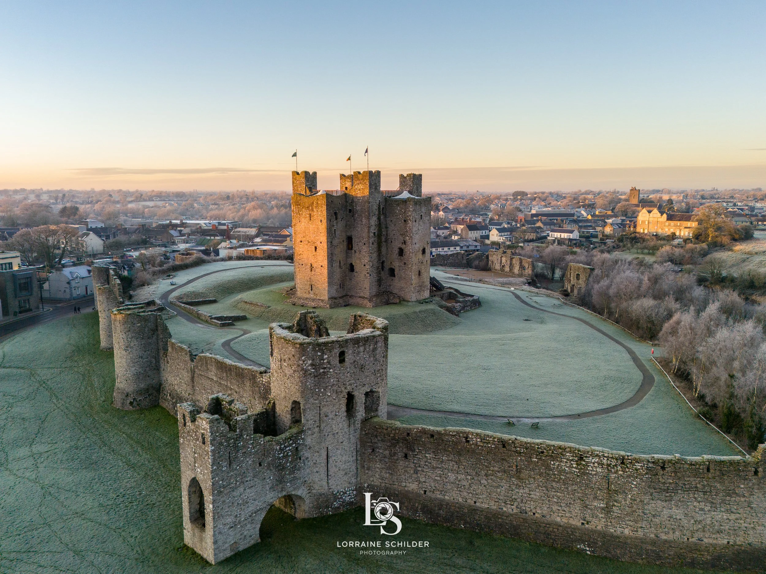Aerial view of Trim castle with stone towers and walls, surrounded by a grassy landscape under a clear sky at sunset.
