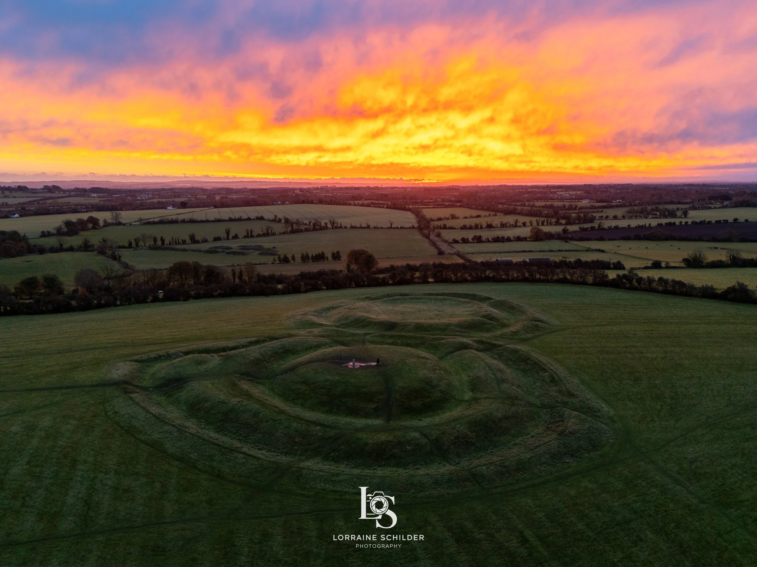 A vibrant sunset over a rural landscape with green fields, trees, and a hill with a statue at its center.