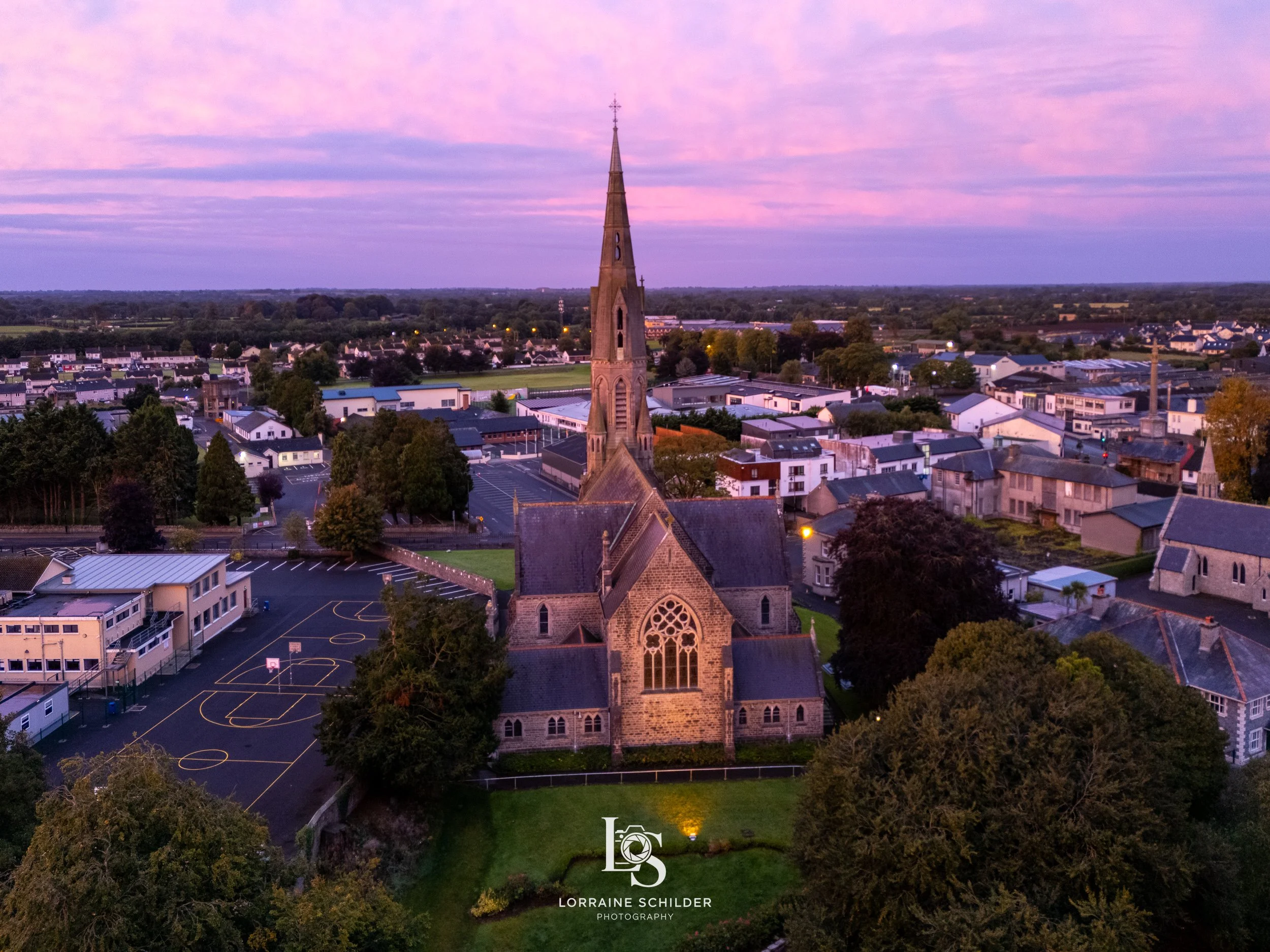 St Patricks Church with a tall steeple surrounded by trees and houses during sunrise with pink and purple sky