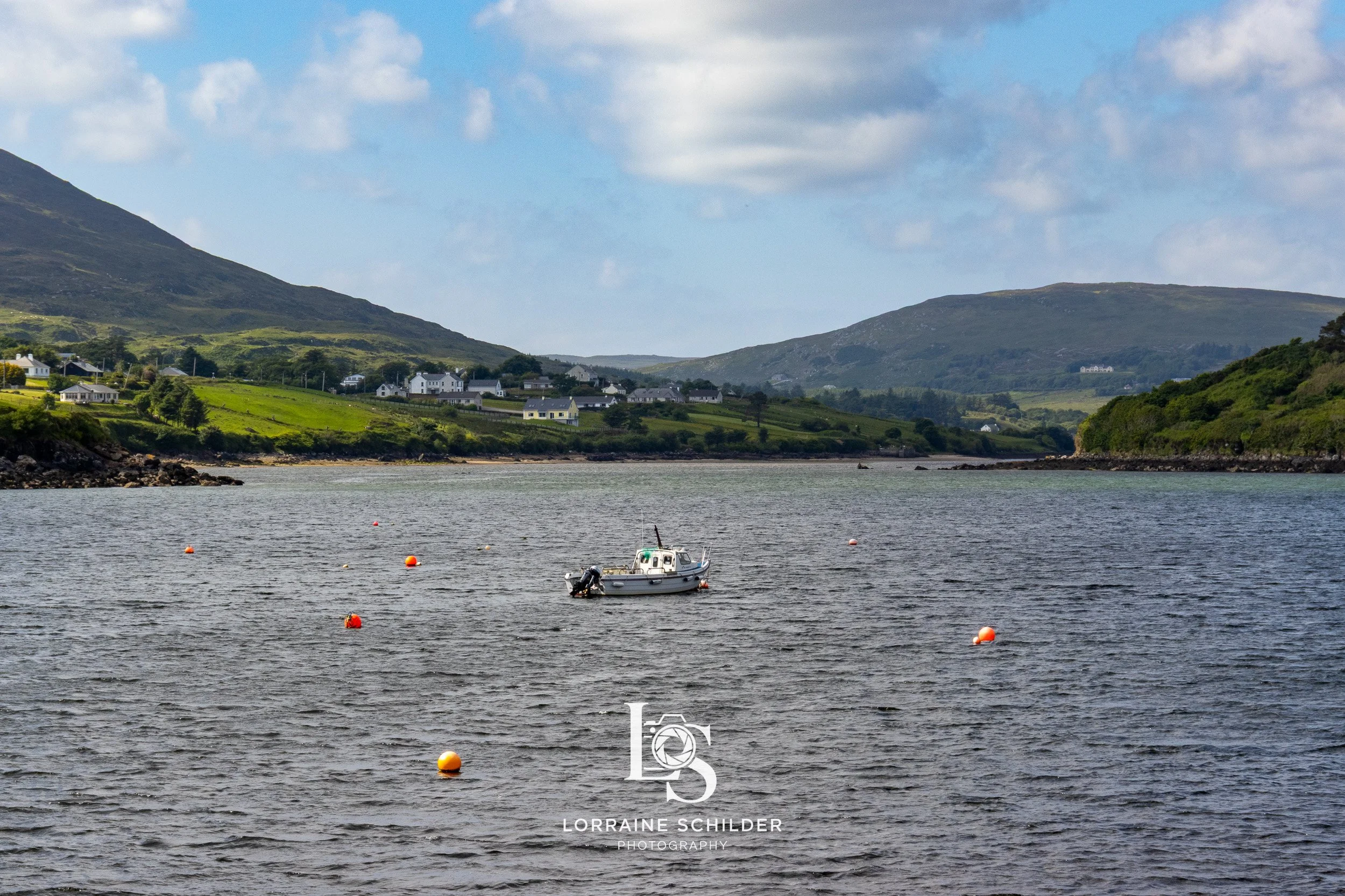 A small boat sailing in a calm river with buoys floating on the water, surrounded by green hills and houses in the background under a partly cloudy sky.  Donegal.