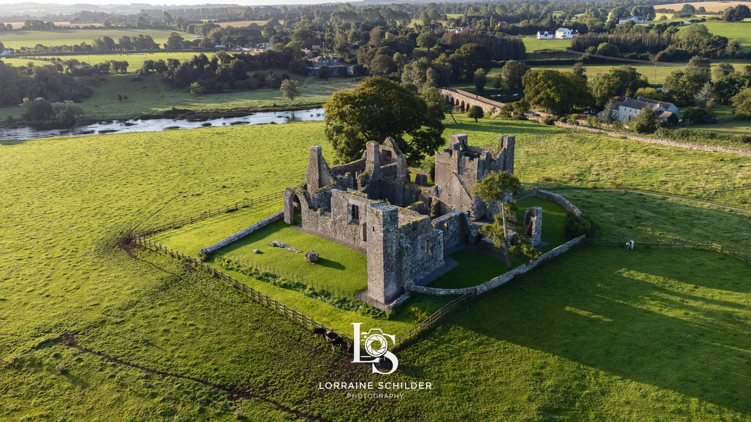 Aerial view of an ancient stone ruin surrounded by green fields, trees, and a fence, with a river and a few houses in the background.  Bective Abbey, Meath.