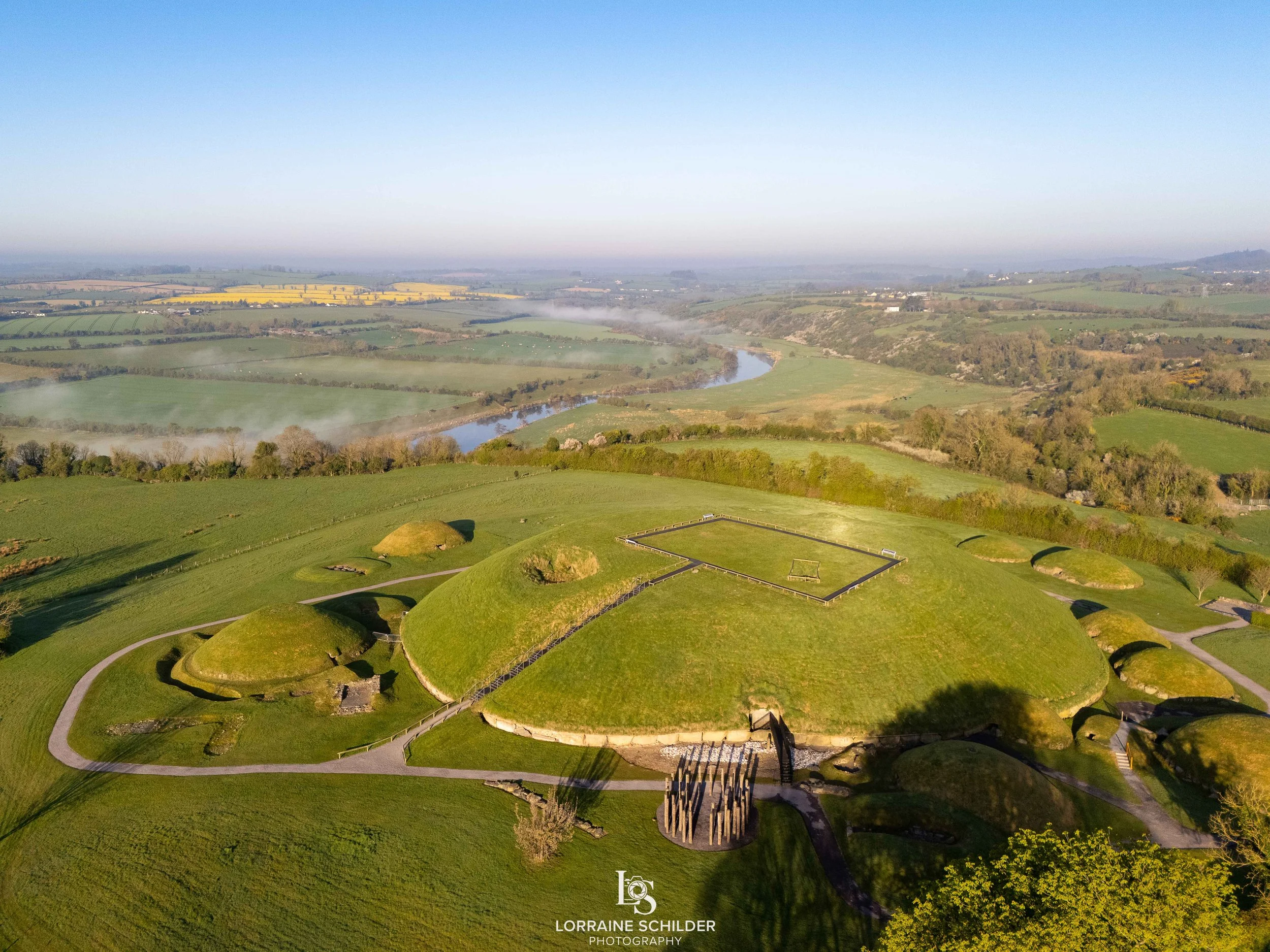 Aerial view of a historic site with mounded earth structures and a rectangular formation on top, surrounded by green grass and pathways, with a river and rolling countryside in the background under a clear sky. Knowth, Meath.