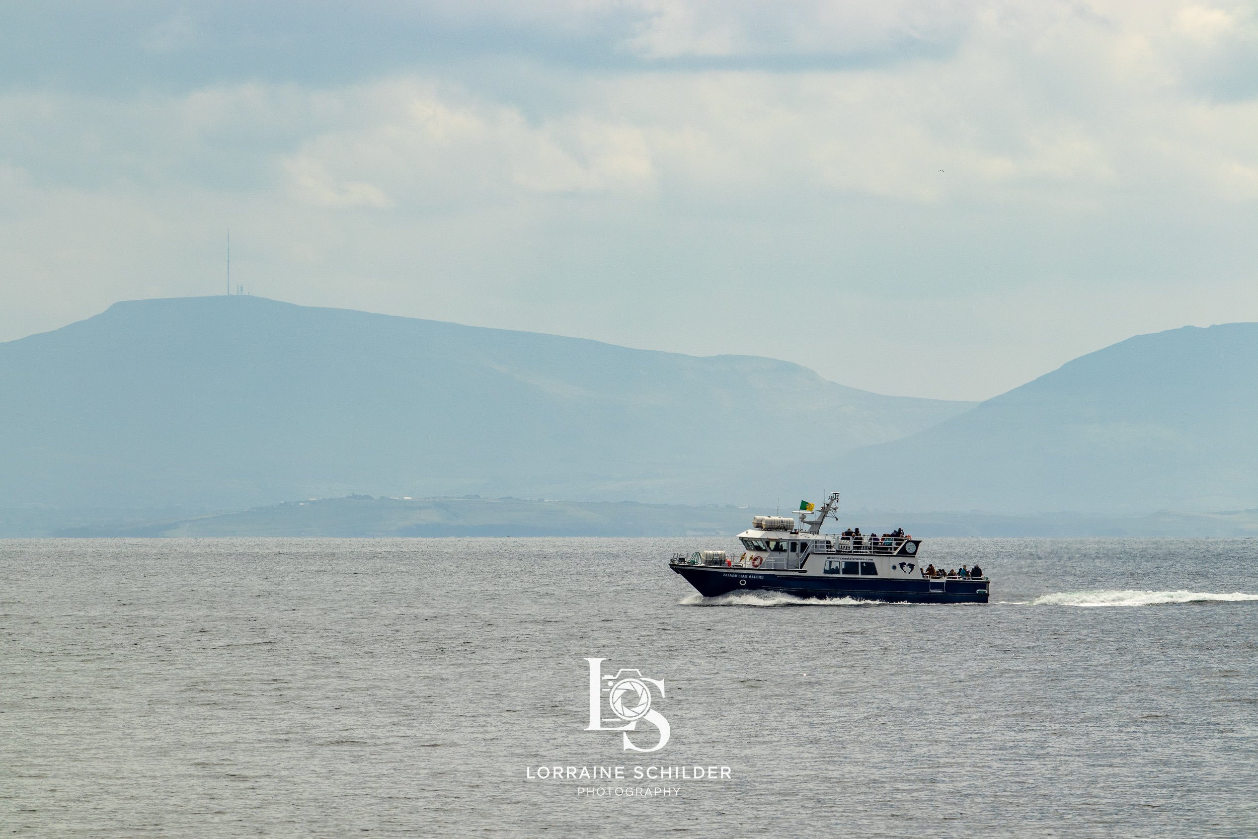 A boat with people on it sailing on a body of water with mountains in the background under a cloudy sky.  Donegal.