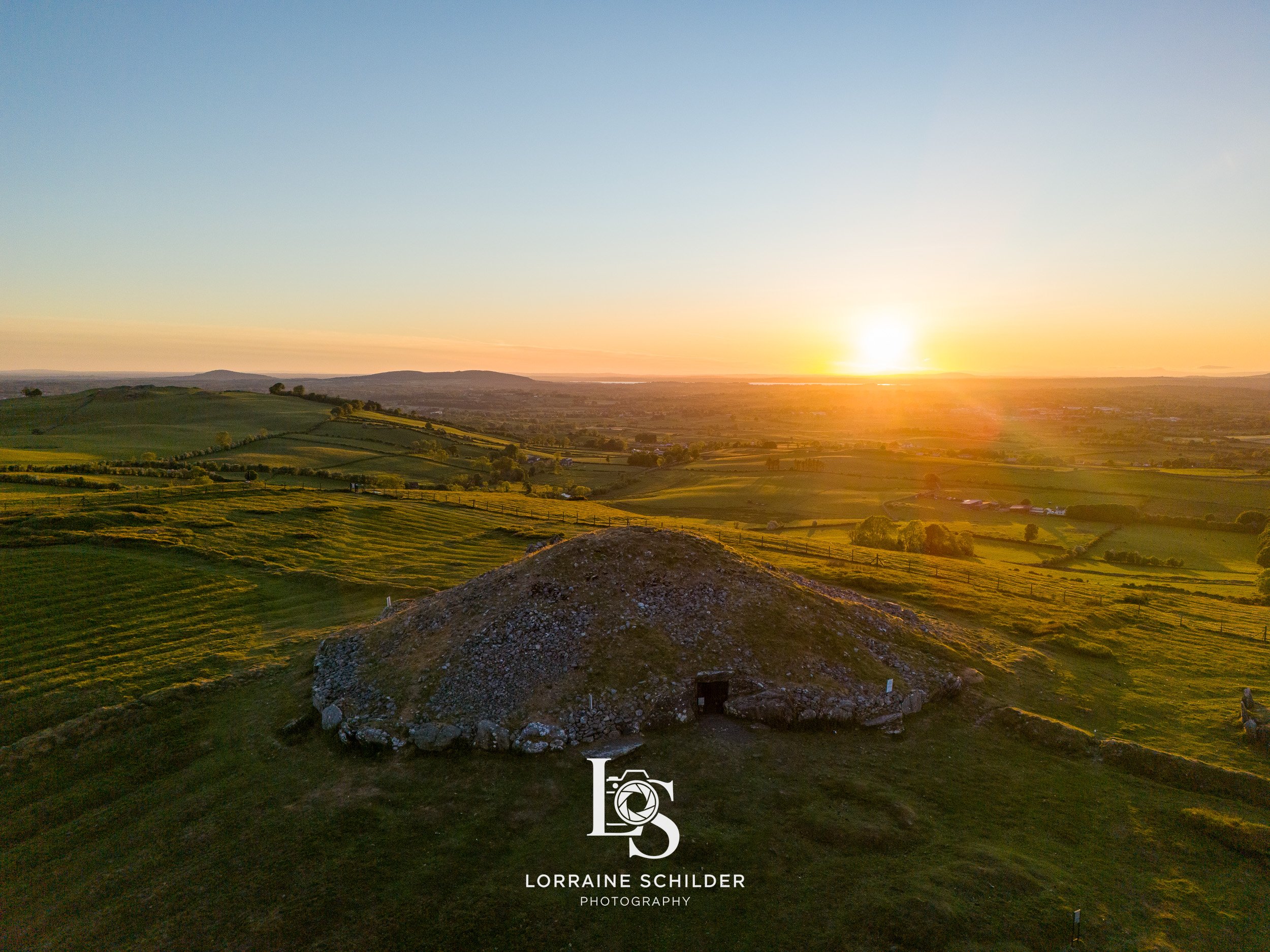 An ancient rounded stone burial mound on a grassy hill at sunset with rolling green fields and distant hills in the background. Loughcrew, Meath.