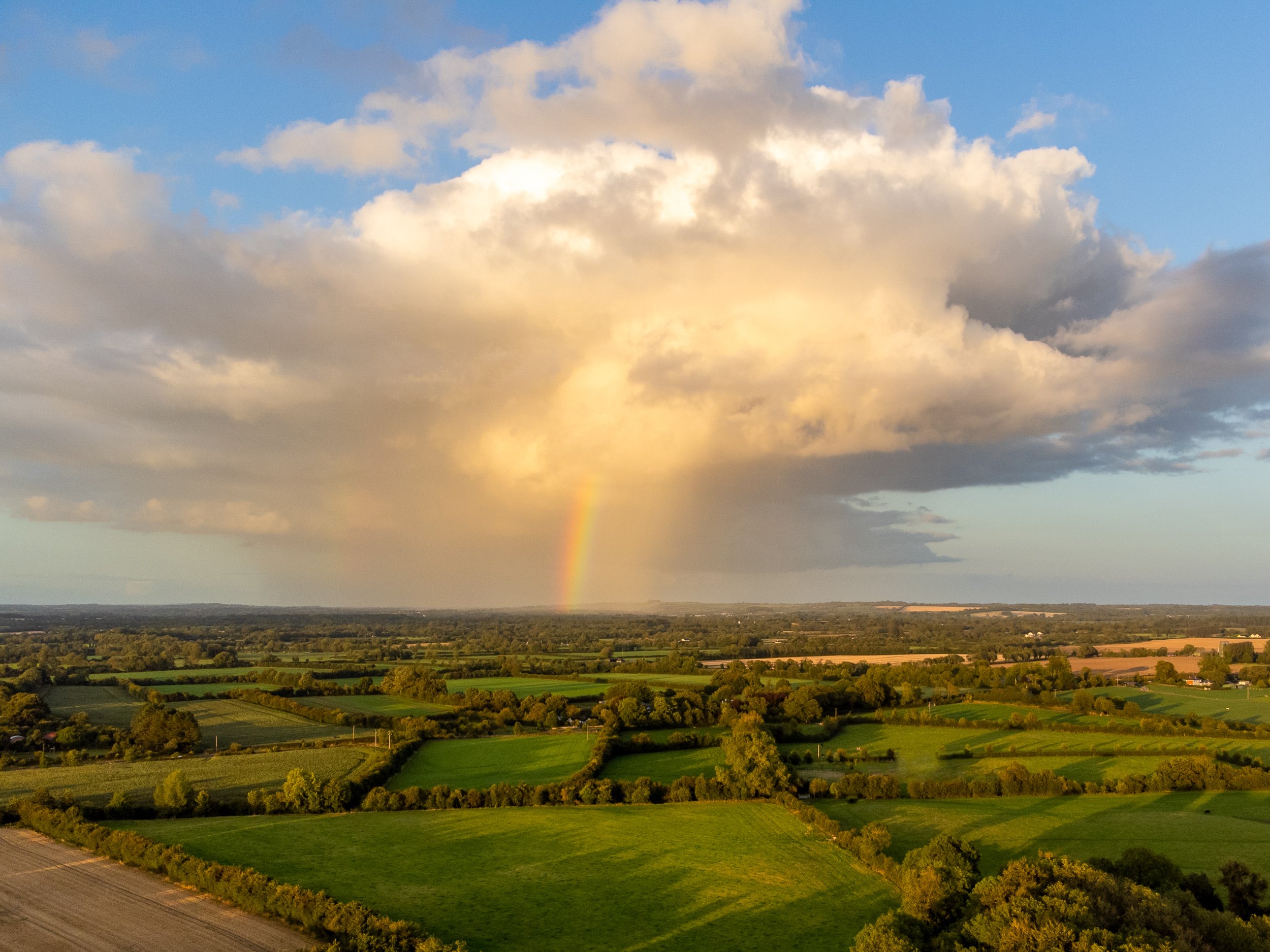 A landscape view of green farmland and trees with a rainbow visible in the distance beneath partly cloudy sky.