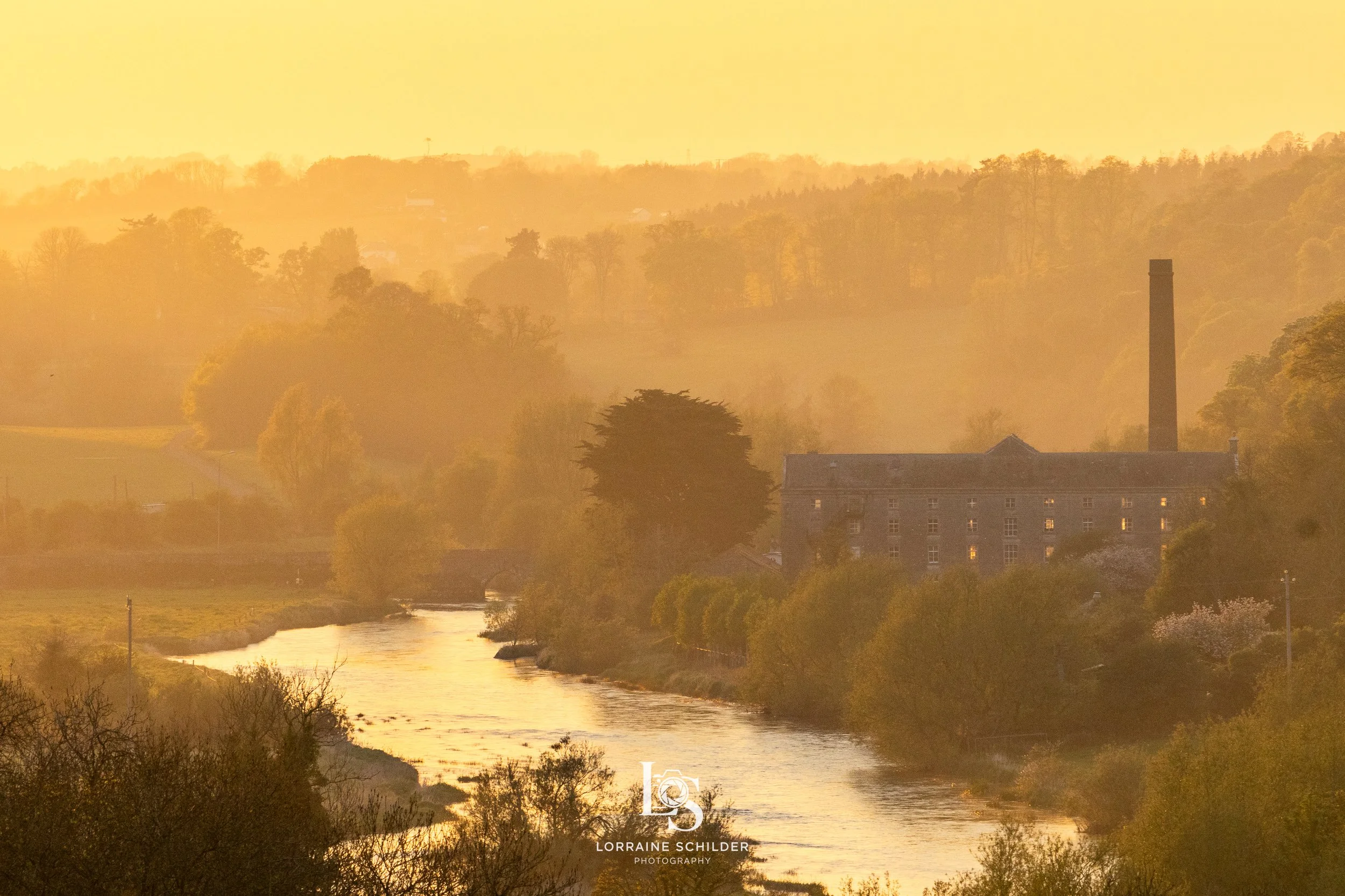 Sunset over a river with trees and an old factory building with a tall chimney in the background.  Slane, Meath.