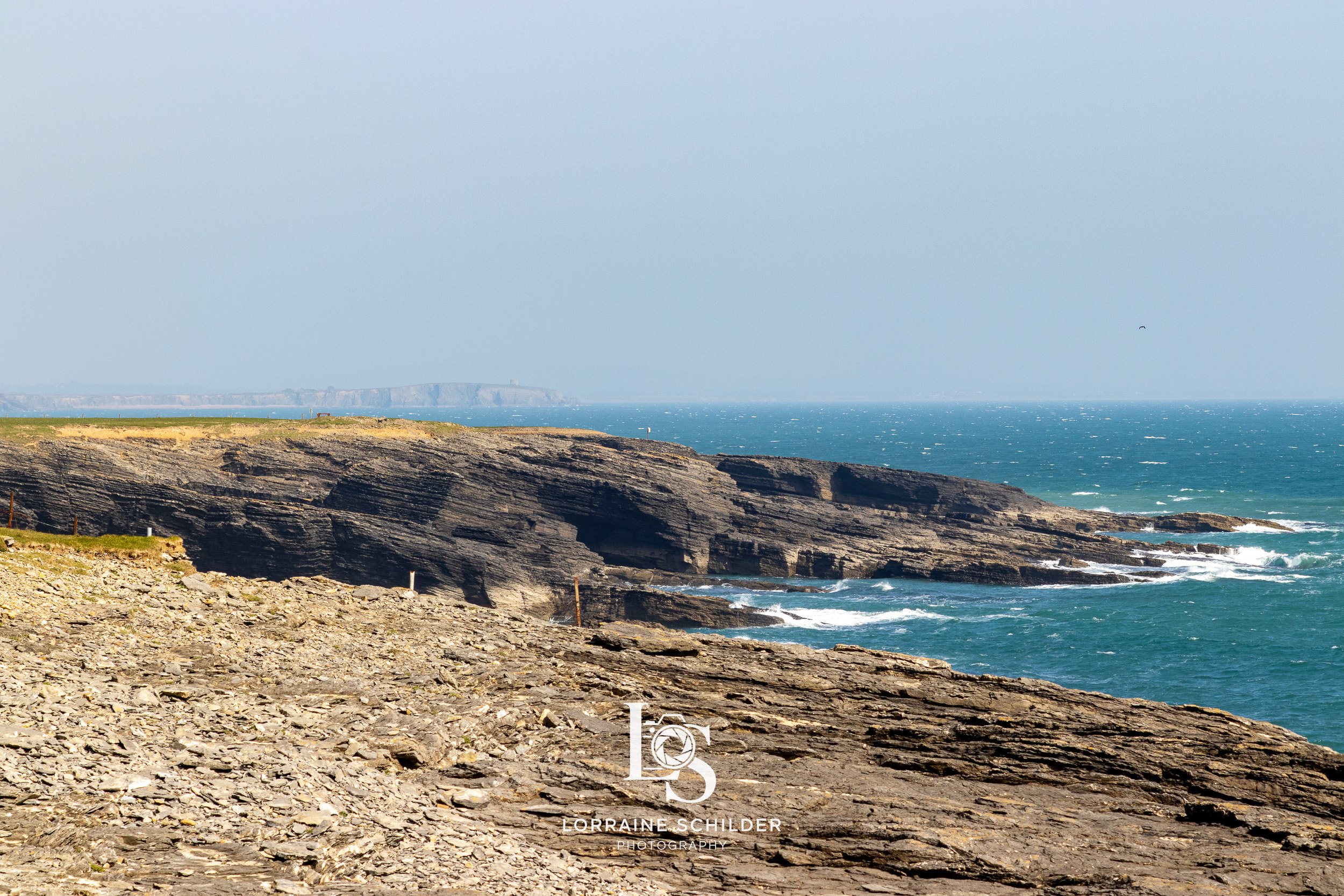 Rocky coastal cliffs with waves crashing below, ocean in the background, and a hazy sky. Wexford.