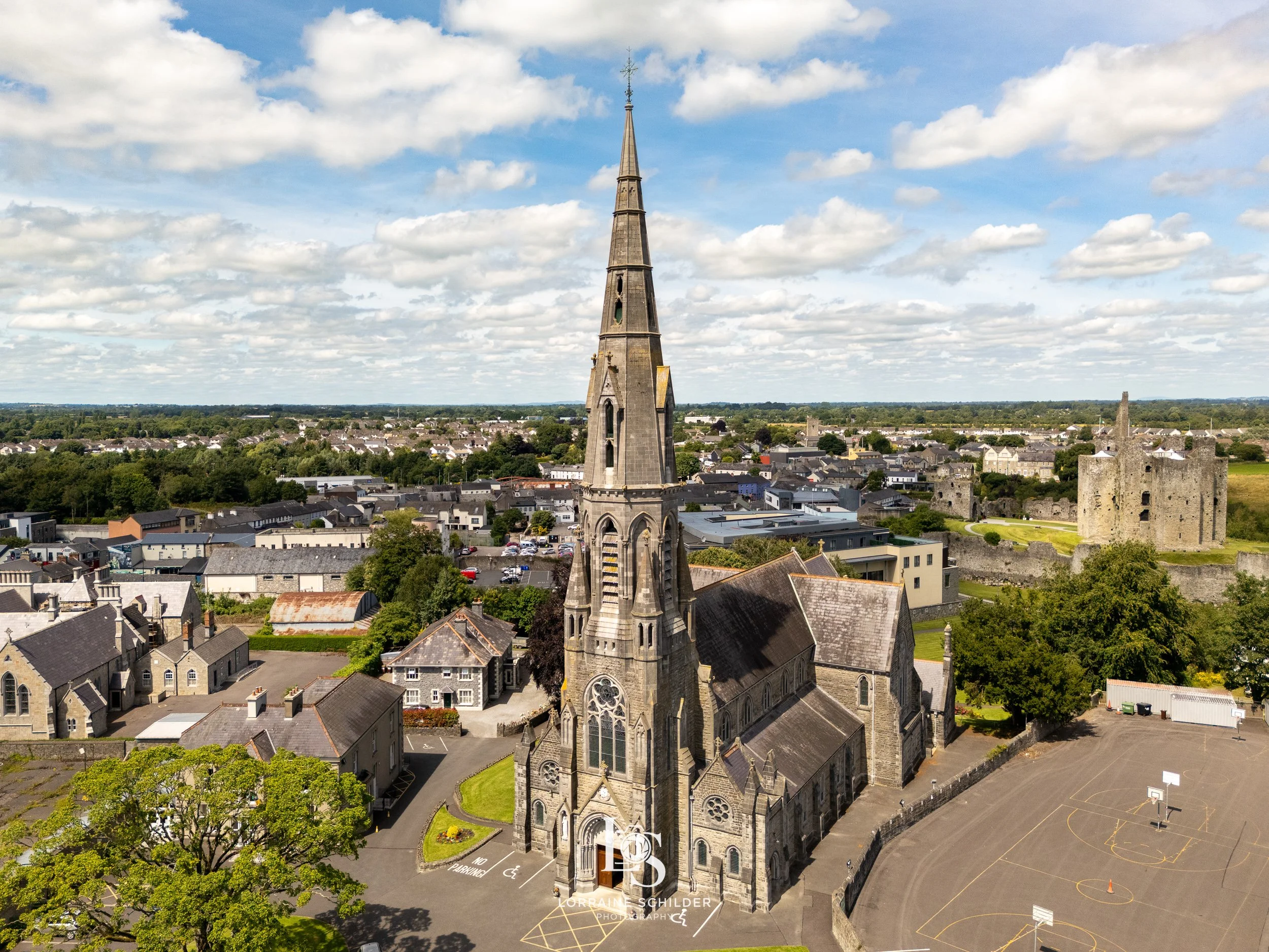 St Patricks church with a tall steeple situated in Trim town, surrounded by residential houses, a Trim castle ruin, and an empty basketball court under a partly cloudy sky.