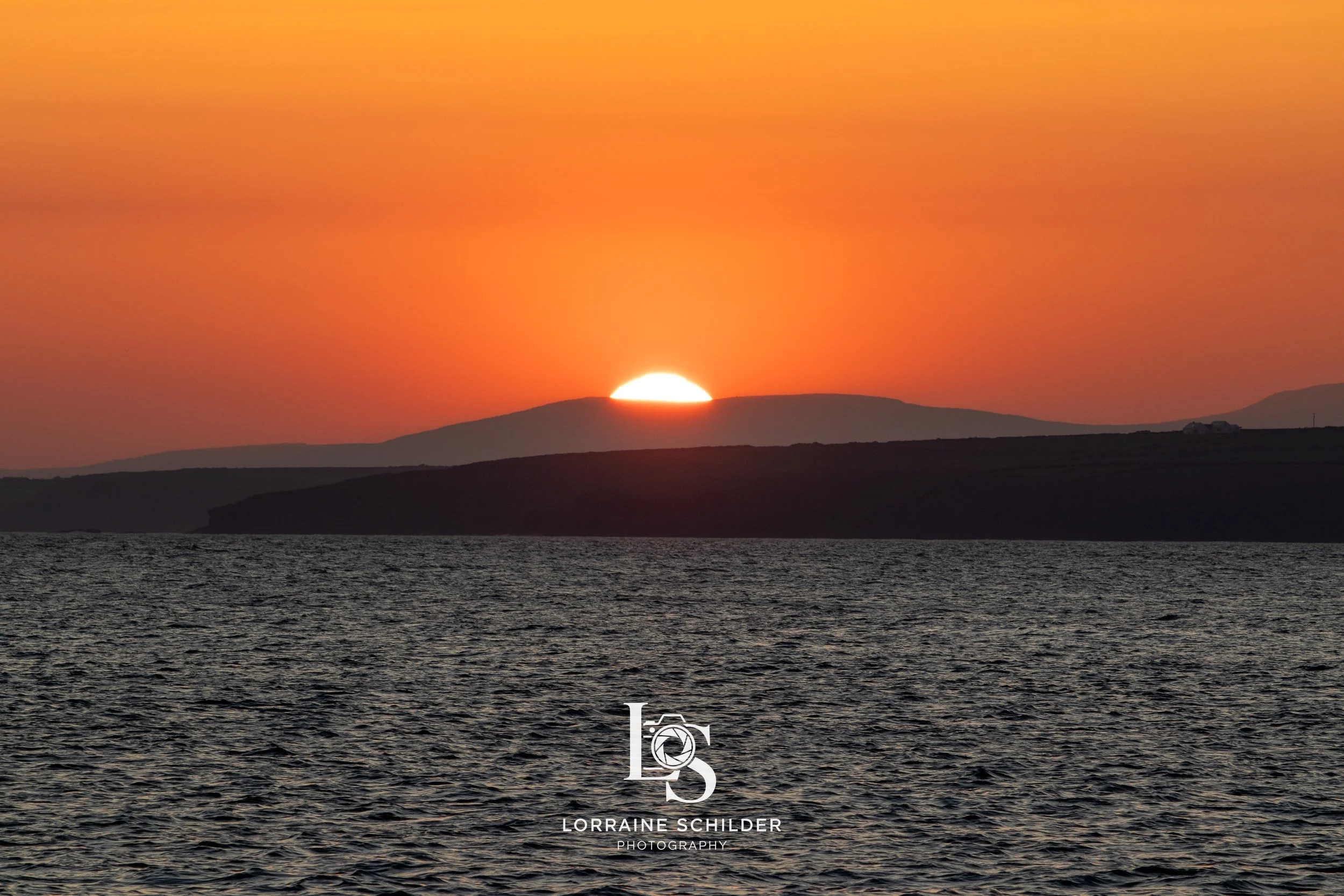 Sun setting over a body of water near distant hills and mountains at dusk with orange sky. Wexford.
