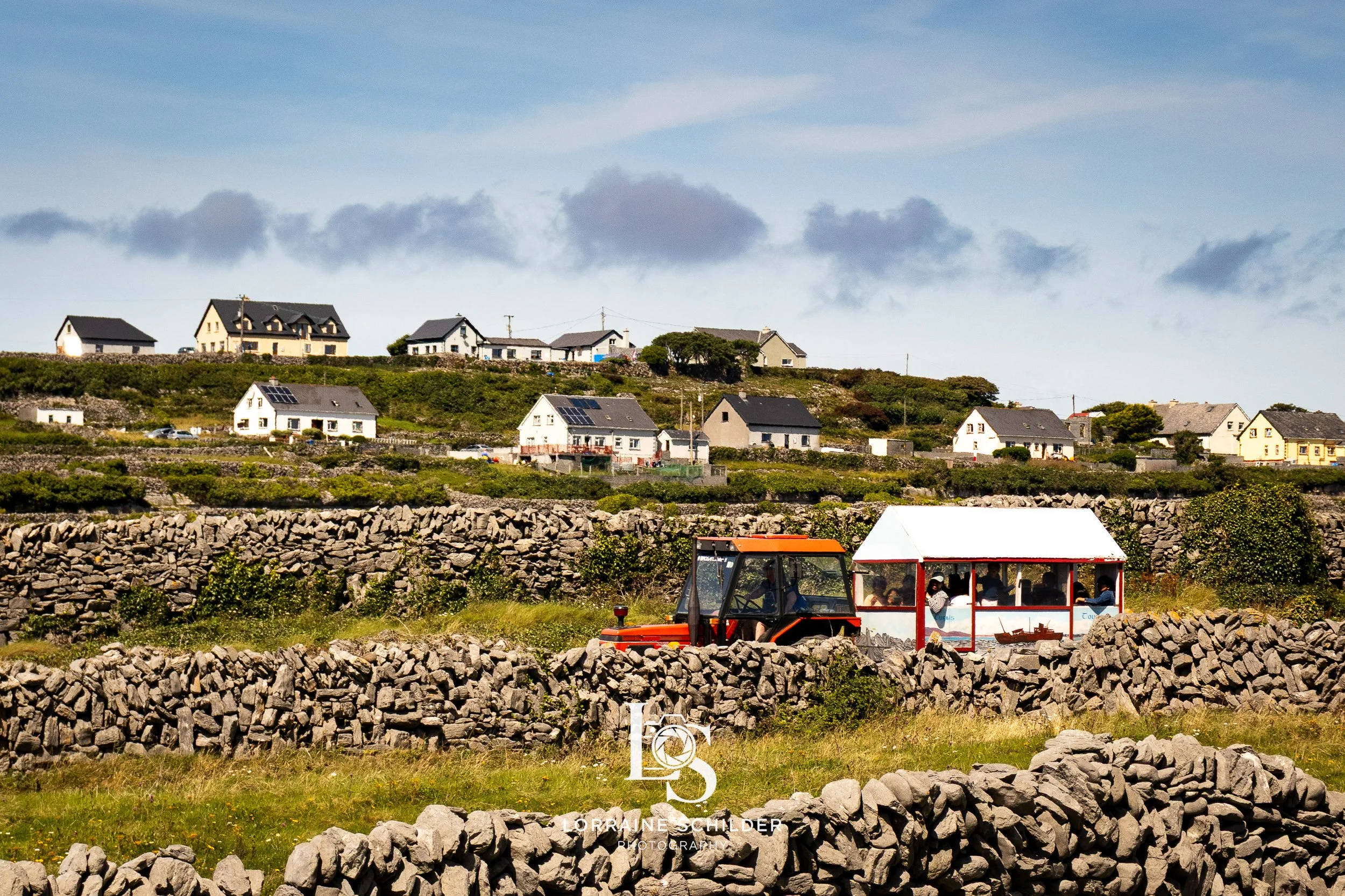 A countryside scene with a tractor pulling a white structure over stone walls, and houses with solar panels on rooftops in the background under a partly cloudy sky.  Inisheer, Galway.