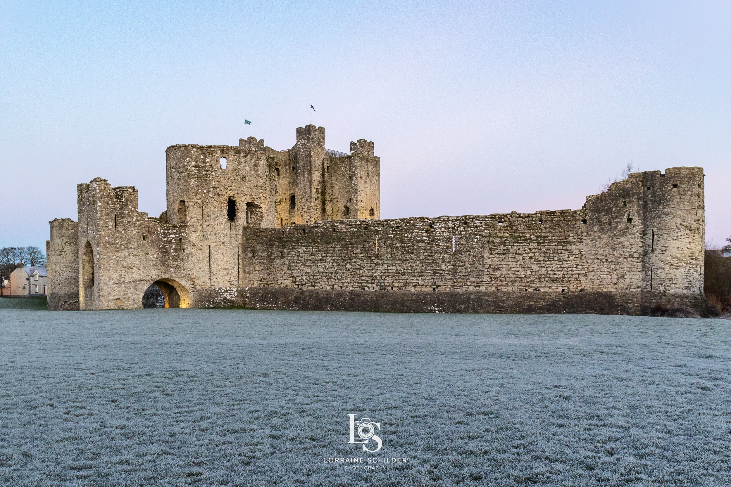 Trim Castle with multiple towers and a surrounding wall, situated on a body of water with a grassy area in the foreground, under a clear sky.