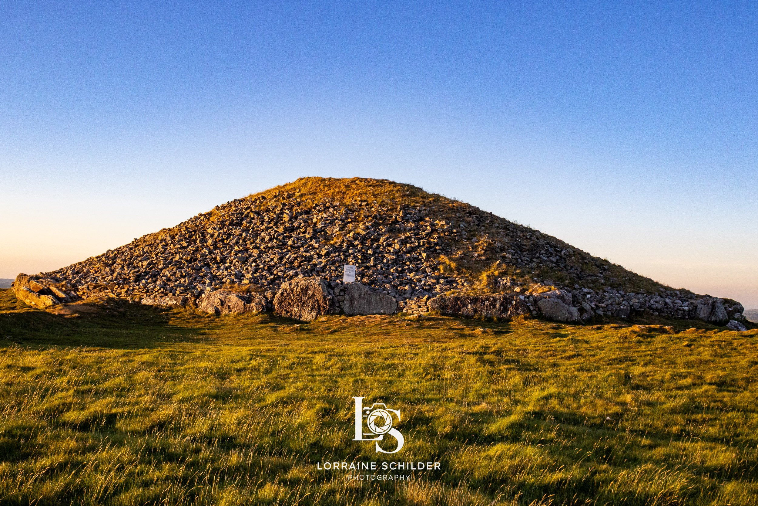 A large grassy field with a hill made of rocks and stones under a clear blue sky during sunset. Loughcrew, Meath.