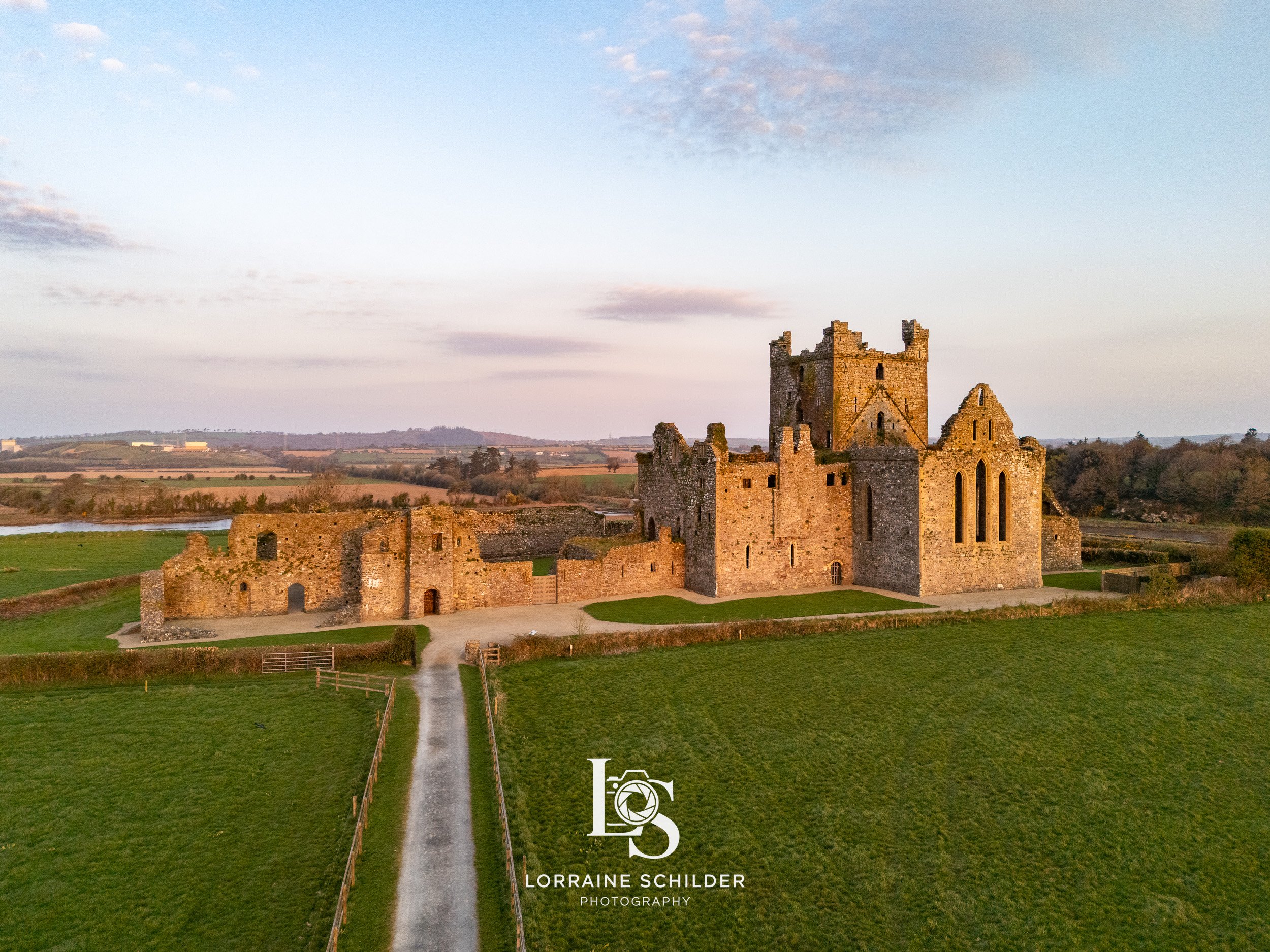 An old stone castle situated on a grassy field with a dirt path leading to its entrance, under a partly cloudy sky during sunrise. Wexford.