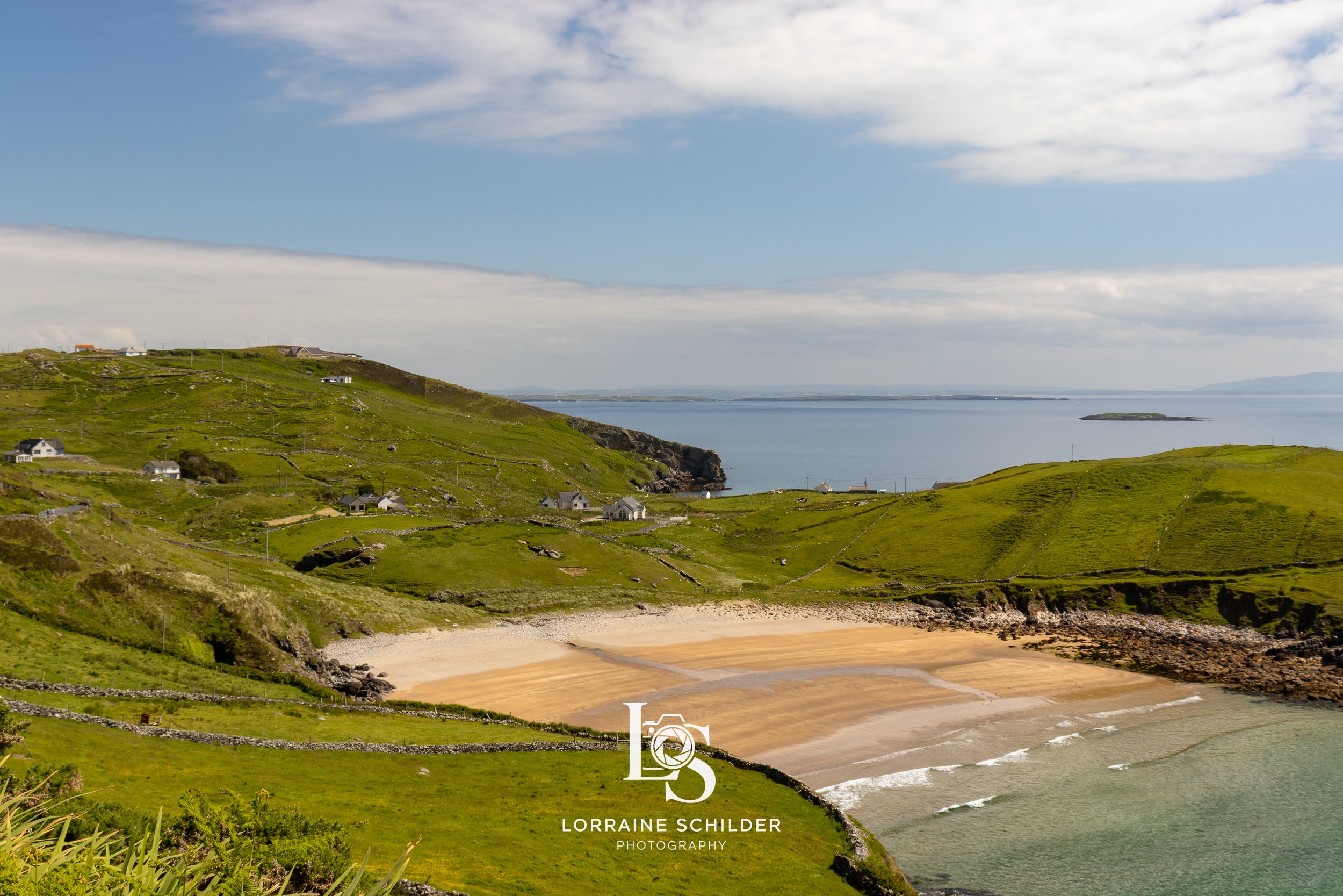 Scenic view of a green hillside with a small sandy beach and waves, overlooking a calm body of water under a partly cloudy sky.  Donegal.