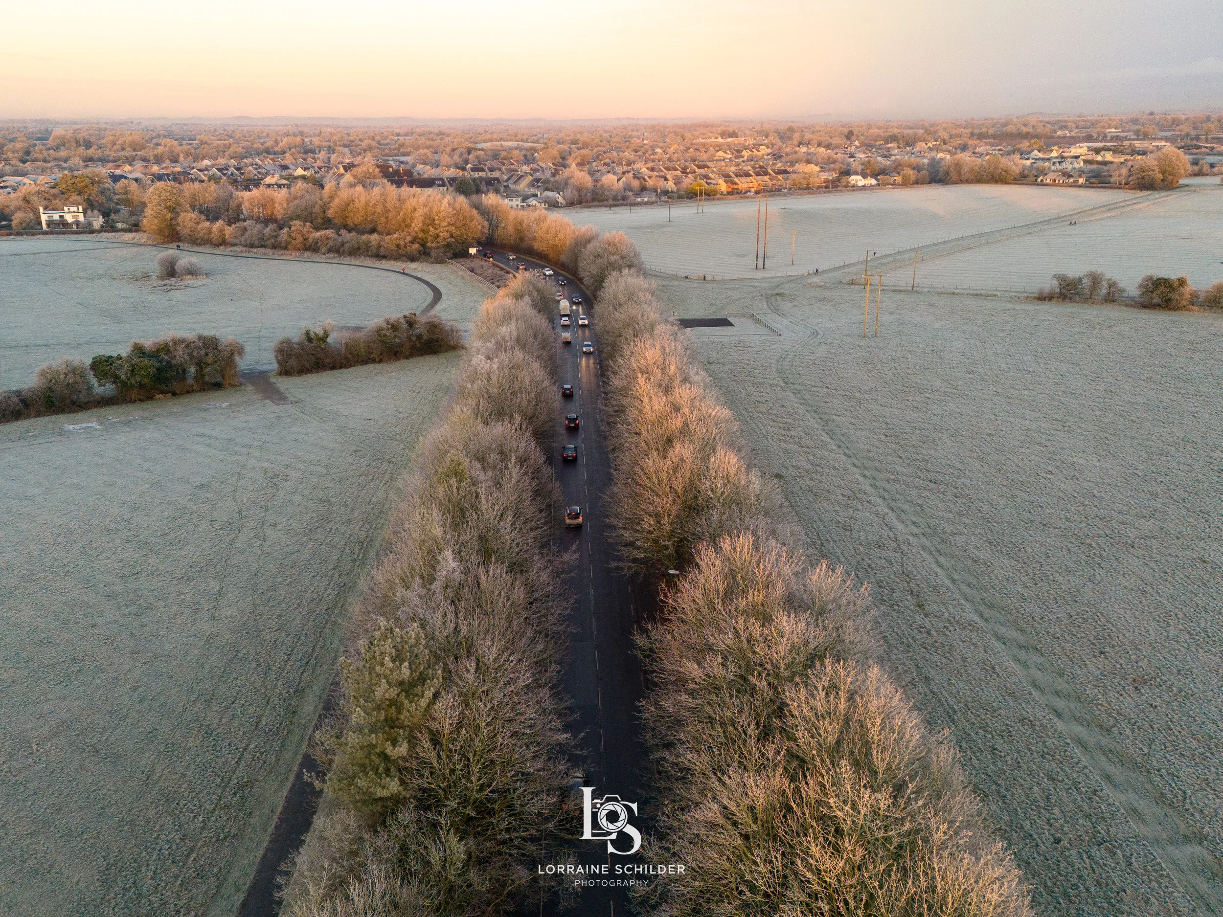 Aerial view of a road with cars driving through a frost-covered landscape at sunset, surrounded by fields, trees, and distant houses. Trim, Meath.