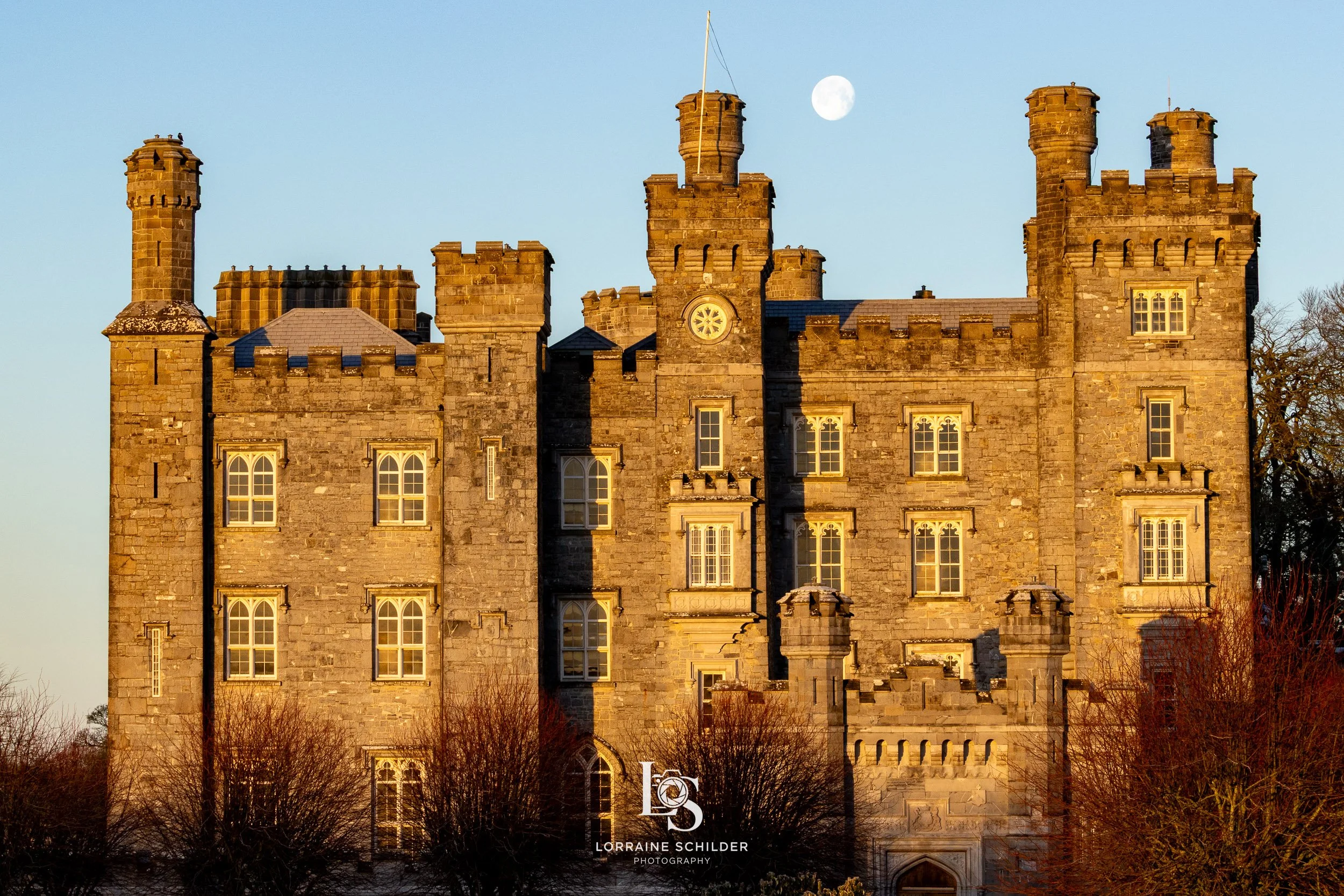 An old stone castle with multiple towers is illuminated by sunlight, with a full moon visible in the sky behind it. Killeen Castle, Meath.