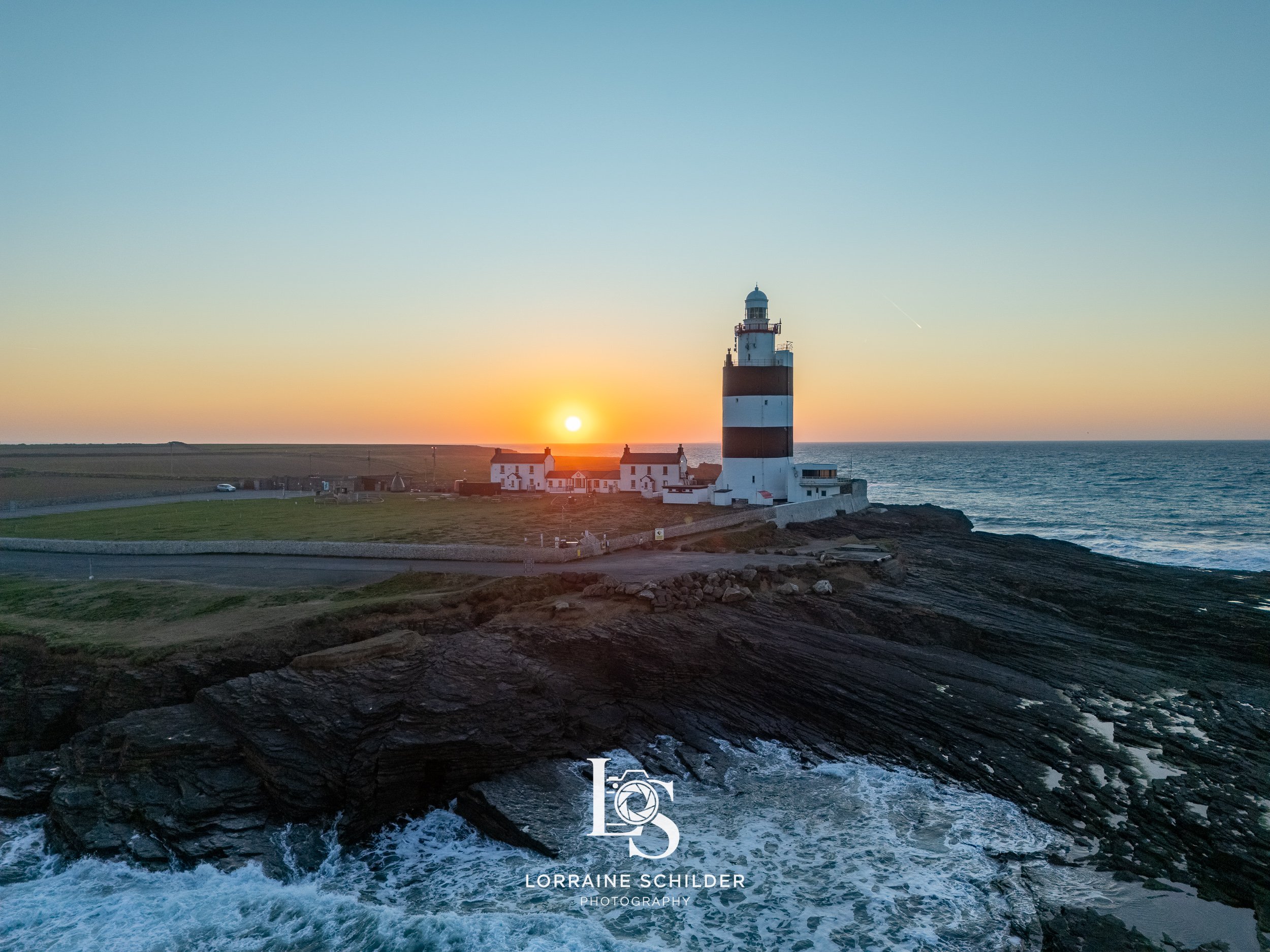 A lighthouse on a rocky coast during sunset, with water crashing against the rocks and a few white houses near the lighthouse. Wexford.