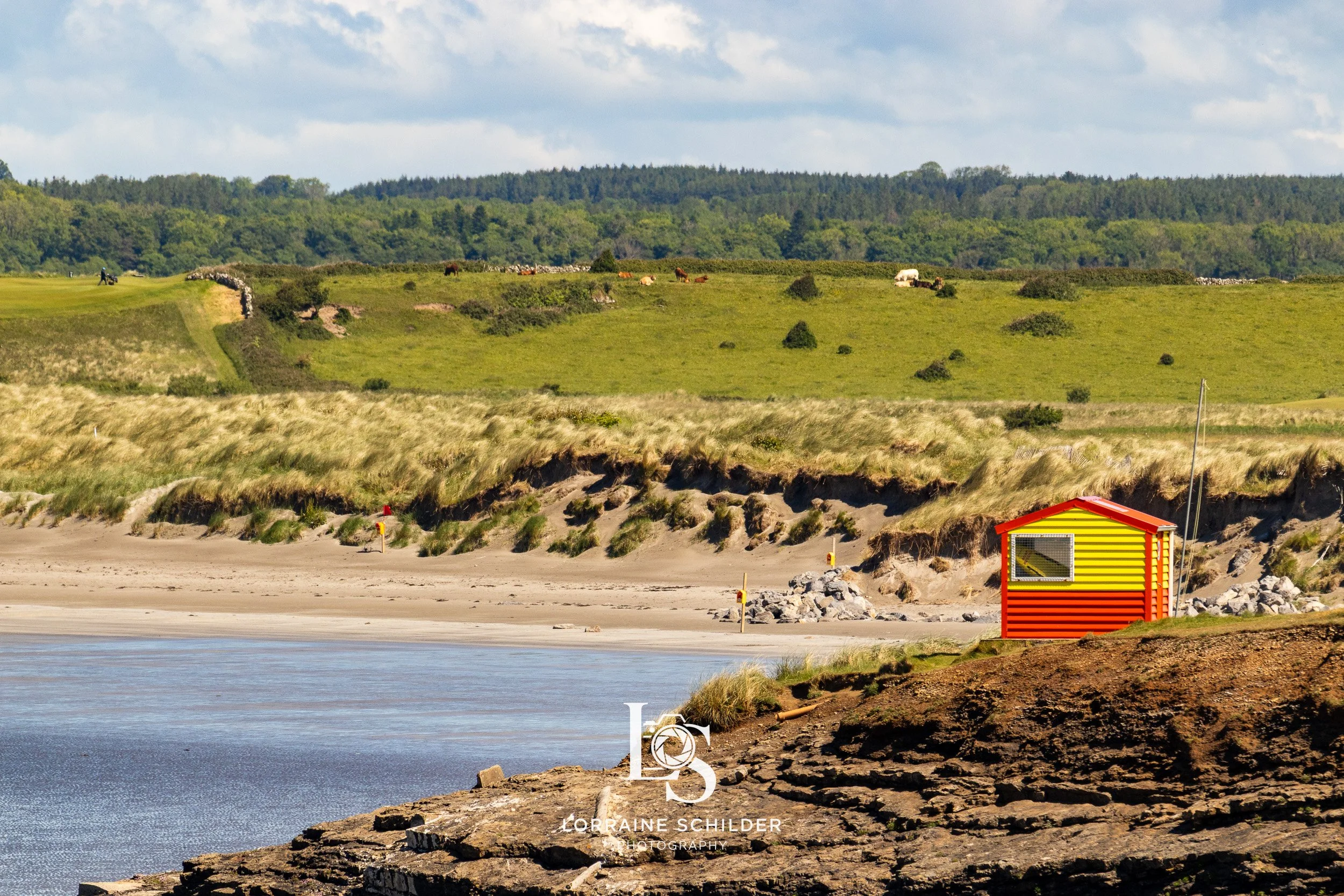 A colorful shoreline with a red, yellow-striped beach hut, sandy beach, grassy dunes, and green hills in the background under a partly cloudy sky. Sligo.
