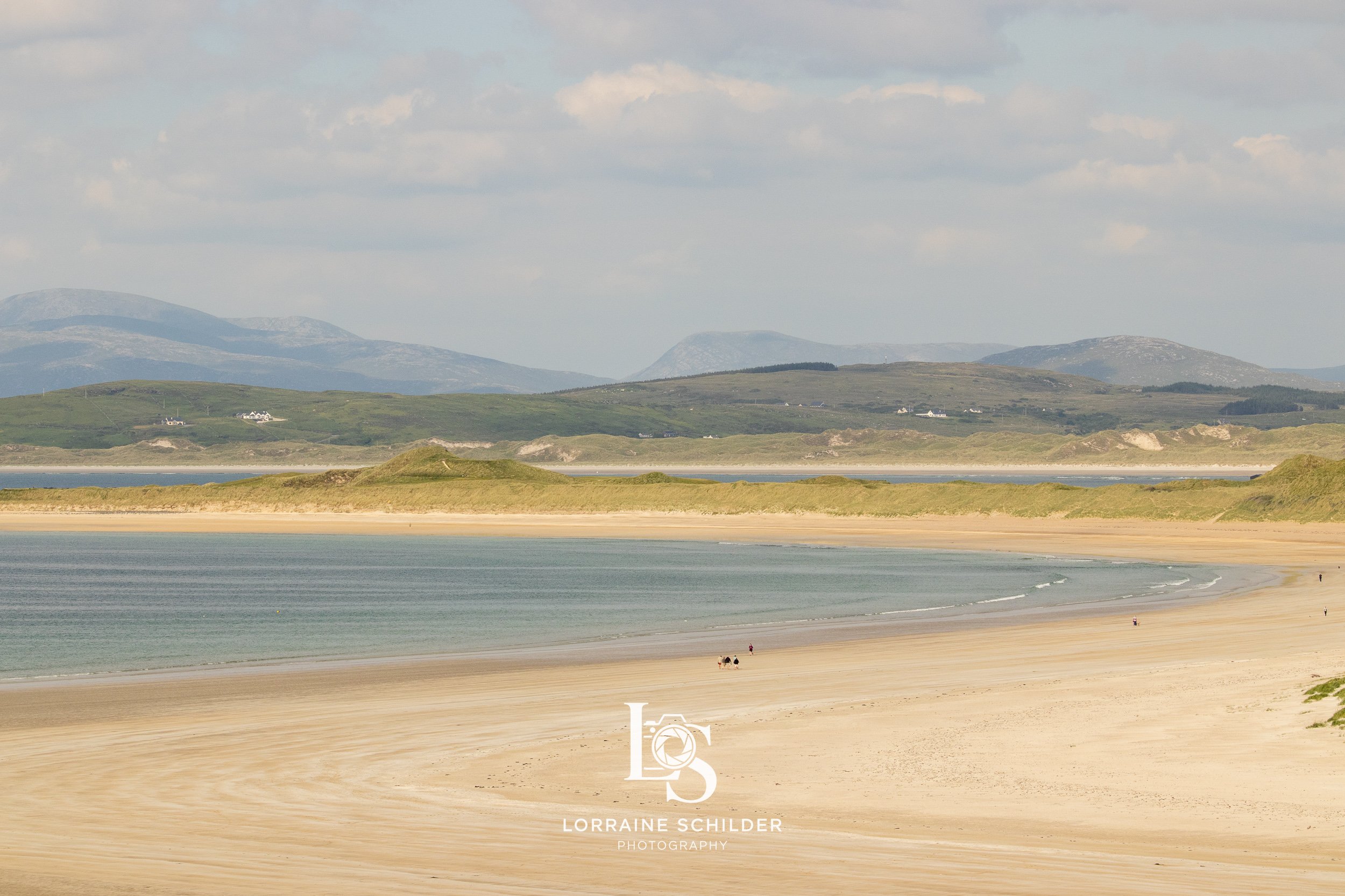 A sandy beach with a few people walking, calm ocean water, and distant green hills under a partly cloudy sky.  Donegal.