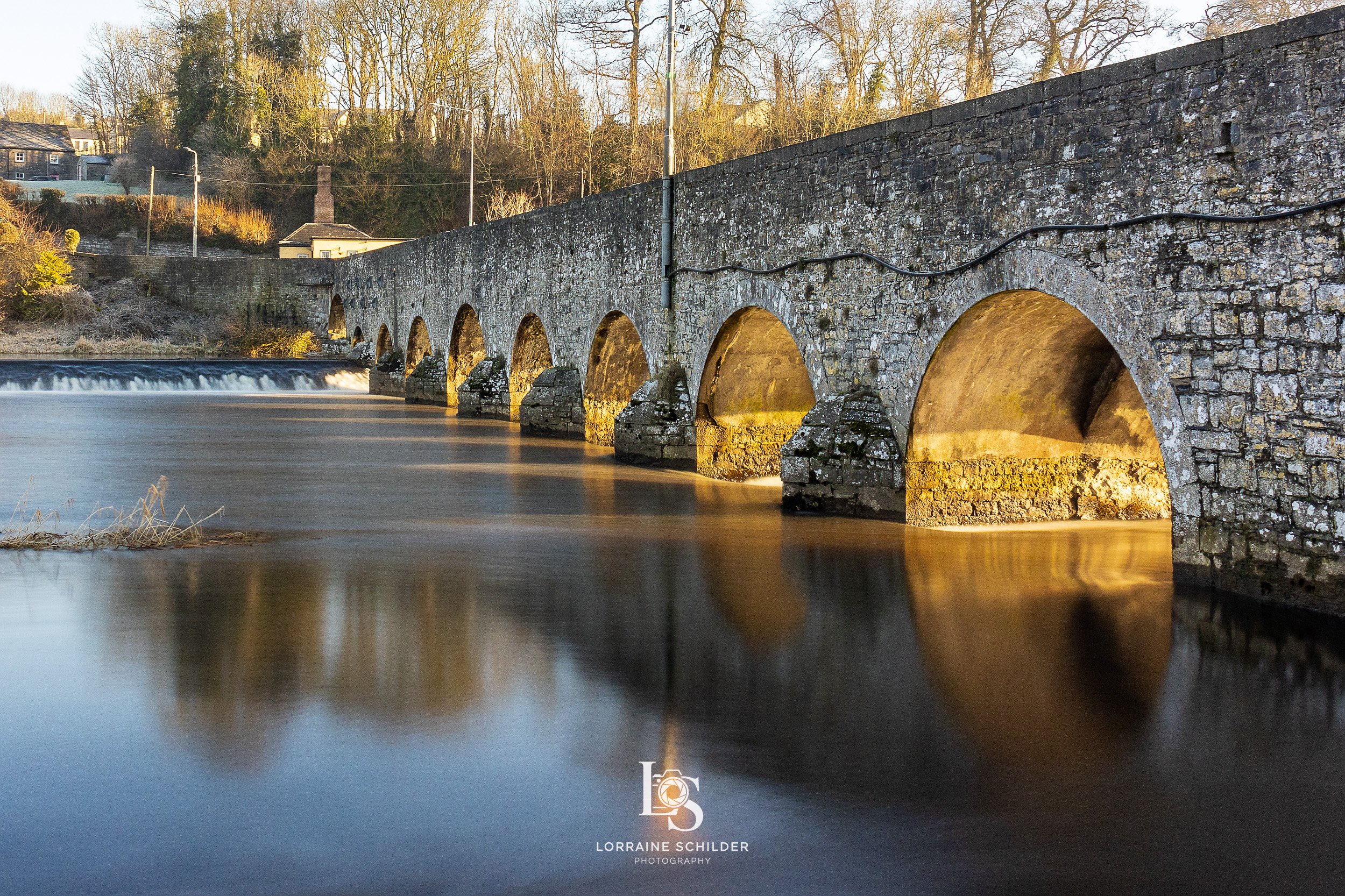 A stone bridge with multiple arches crossing a river, with a small waterfall on one side, during golden hour. Trees and houses are in the background.  Slane, Meath.