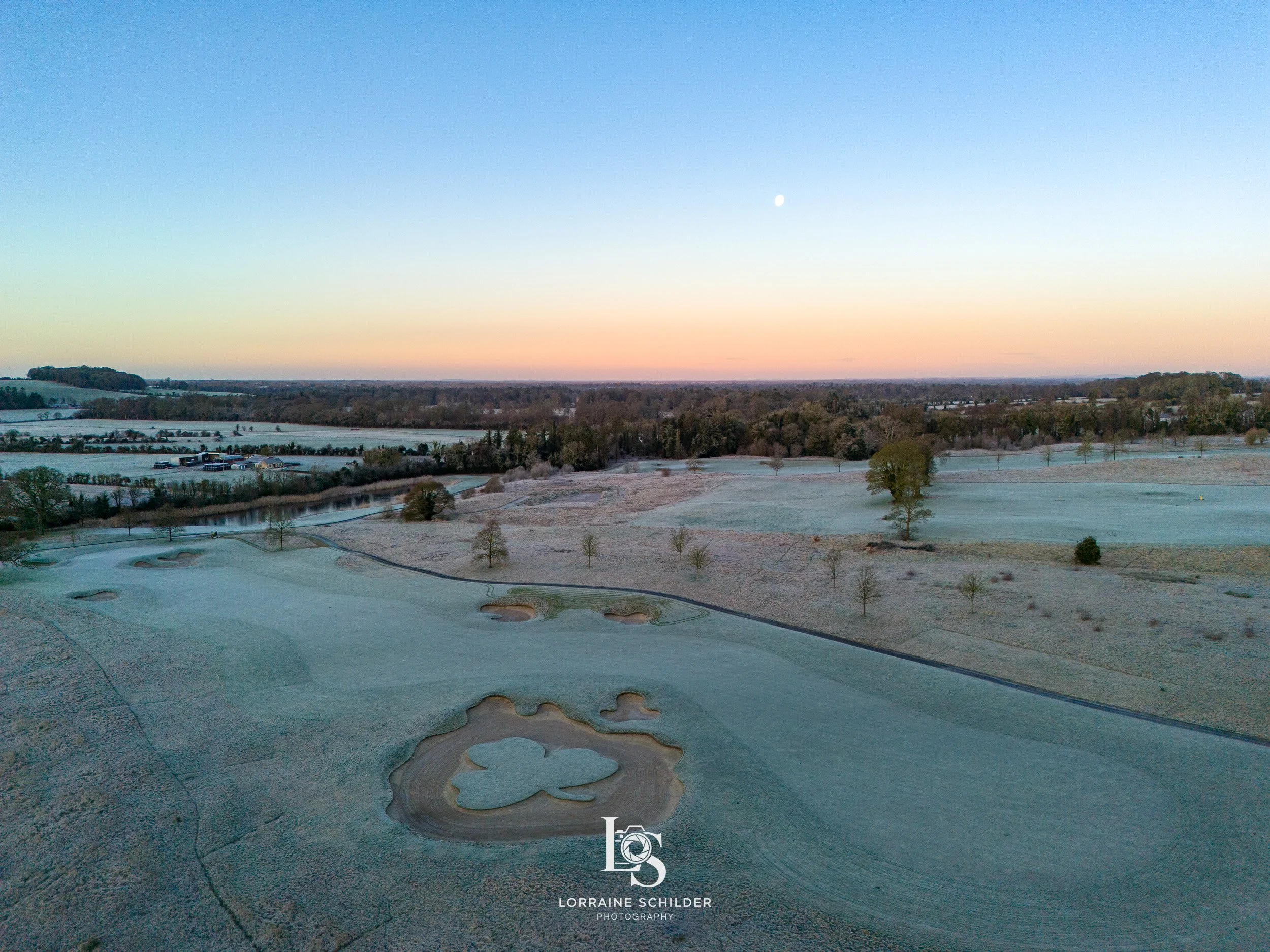 A golf course with sand traps and green grass, trees, and a small pond under a sky with a setting or rising sun and visible moon. Killeen Castle, Meath.