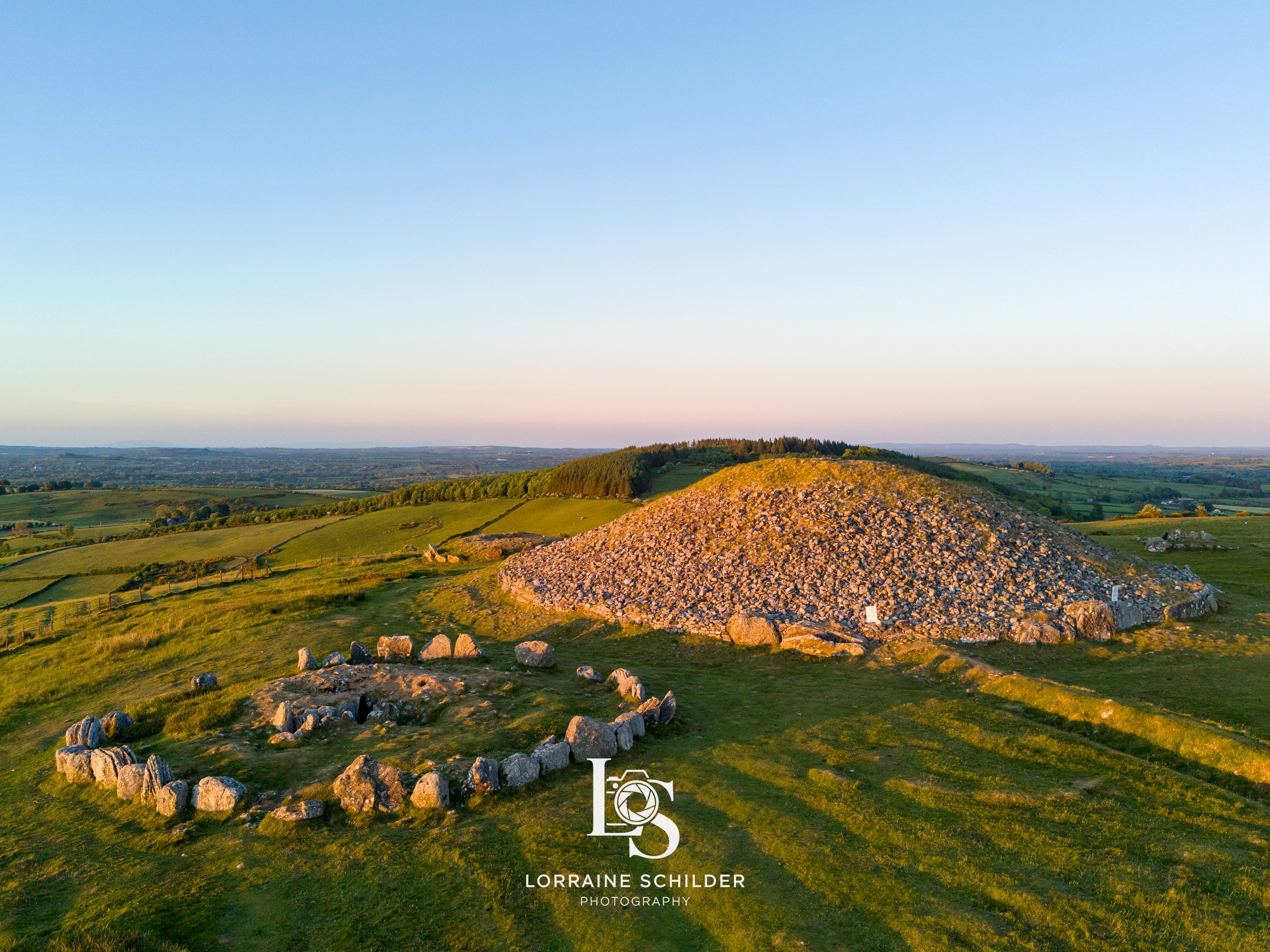 Aerial view of a large ancient stone burial mound or cairn on a grassy hilltop under a clear sky during sunset. Loughcrew, Meath.