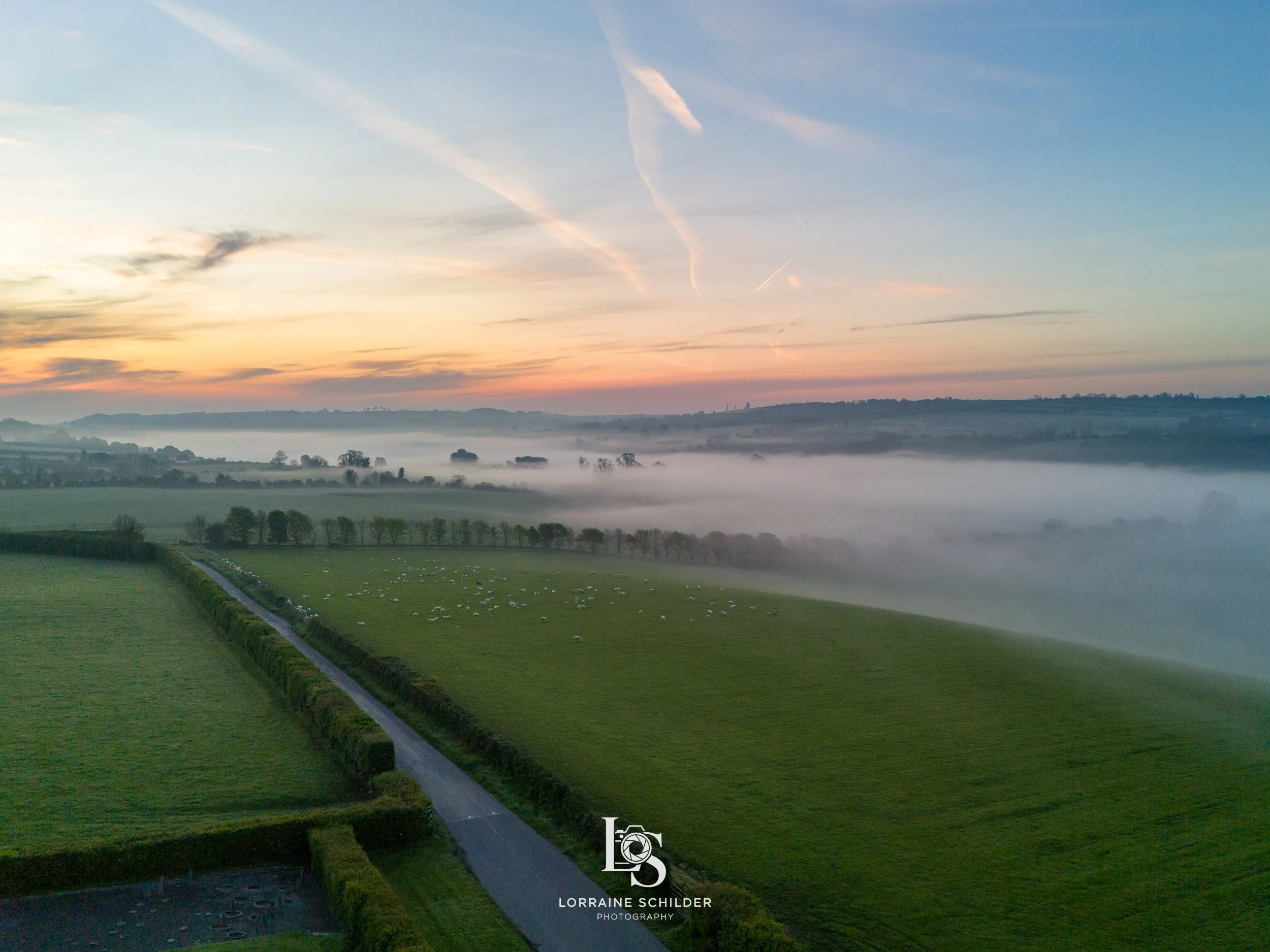 An expansive view of a green landscape at sunrise with a road running through lush fields, mist covering the distant hills, and the sky painted with pastel colors and cloud streaks.  Newgrange, Meath.