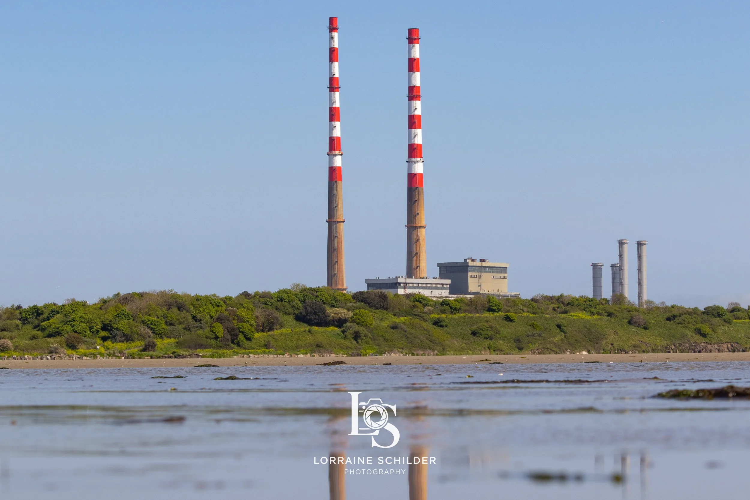 An industrial power plant with tall smokestacks, some painted with red and white stripes, sits on a lush, green landscape by a body of water, under a clear blue sky. Poolbeg, Dublin.