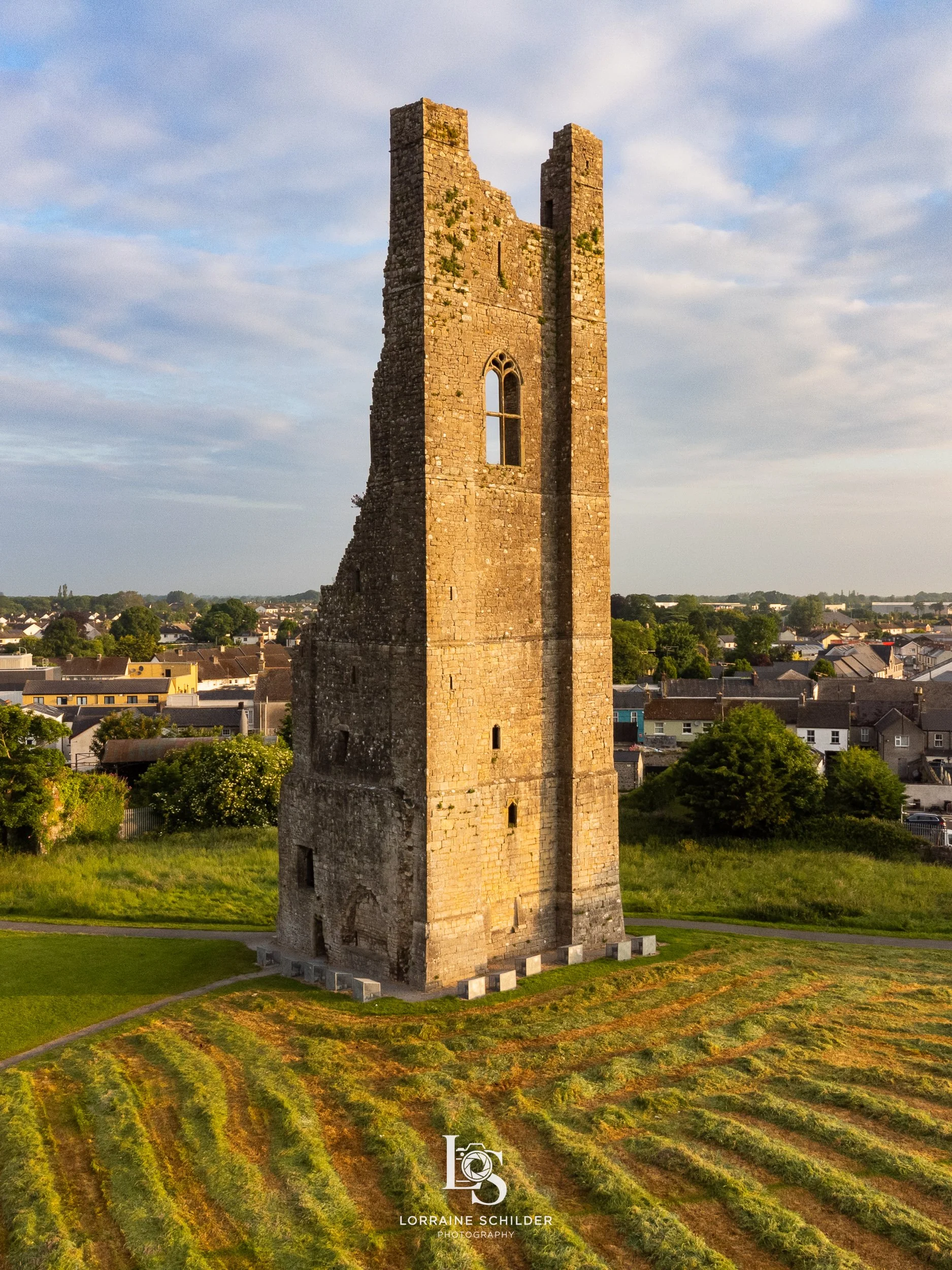 An ancient stone tower stands alone on a grassy hill with a nearby town in the background under a partly cloudy sky during sunrise.