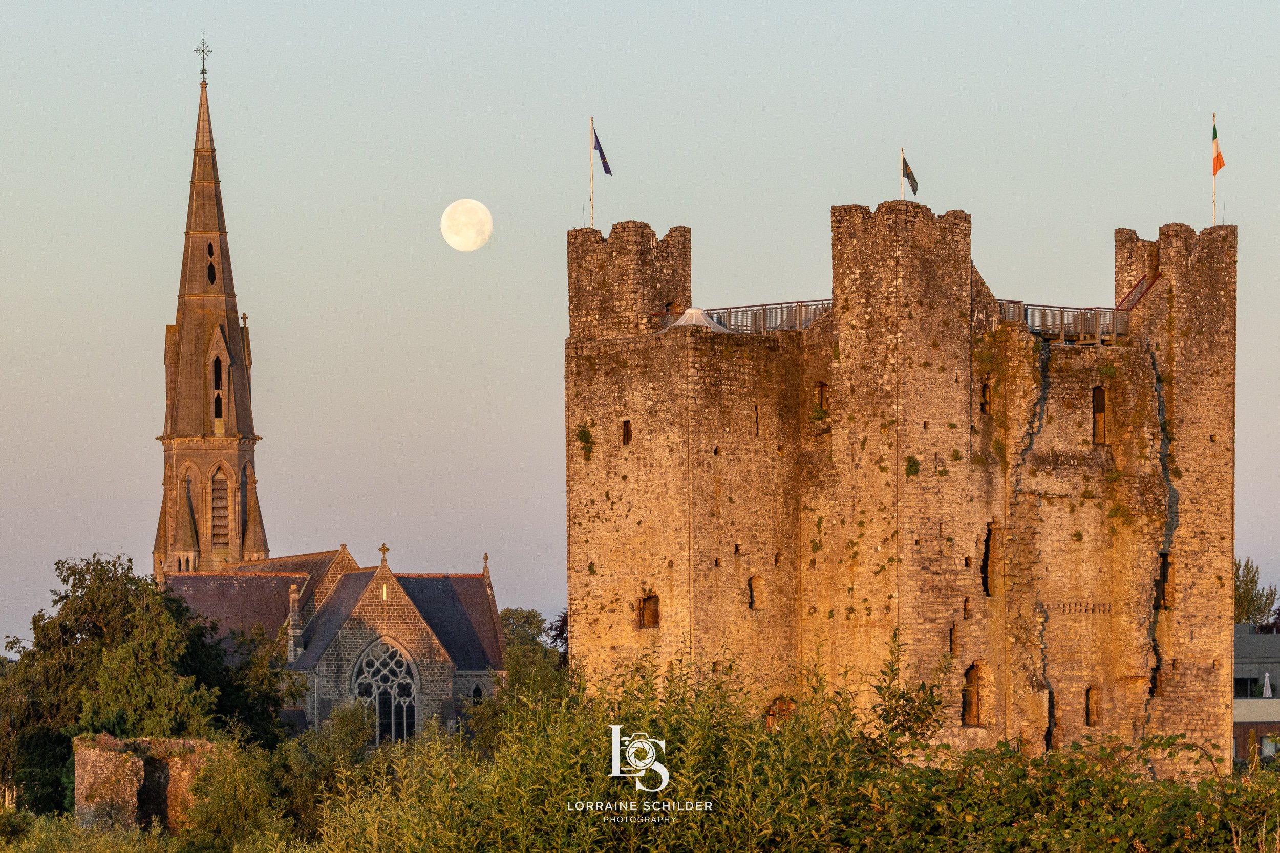 Trim castle with a tall, square tower and flags on top is in the foreground. Behind it, there is St Patricks church with a pointed steeple. The moon is visible in the sky, and the scene is bathed in warm early morning light.