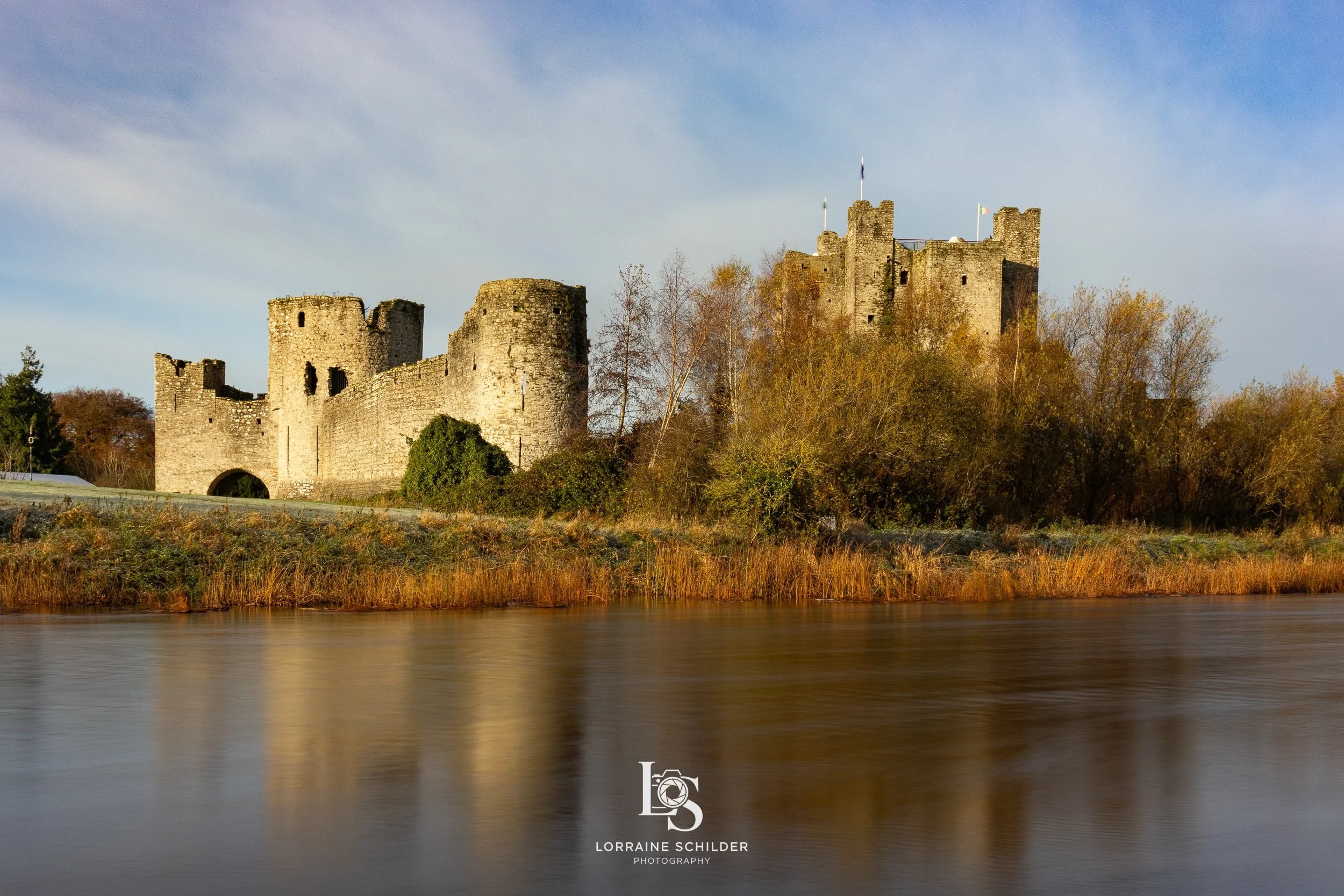 Trim castle on the riverbank with autumn trees and calm water, under a partly cloudy sky.