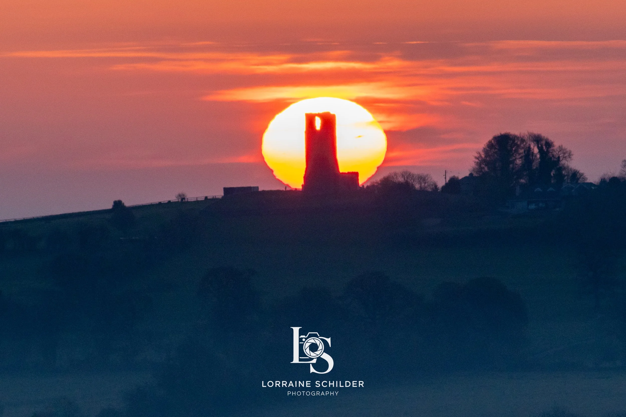 Sunset over a landscape with a silhouetted castle or tower on a hill, with trees and structures in the background. Skyrne, Meath.
