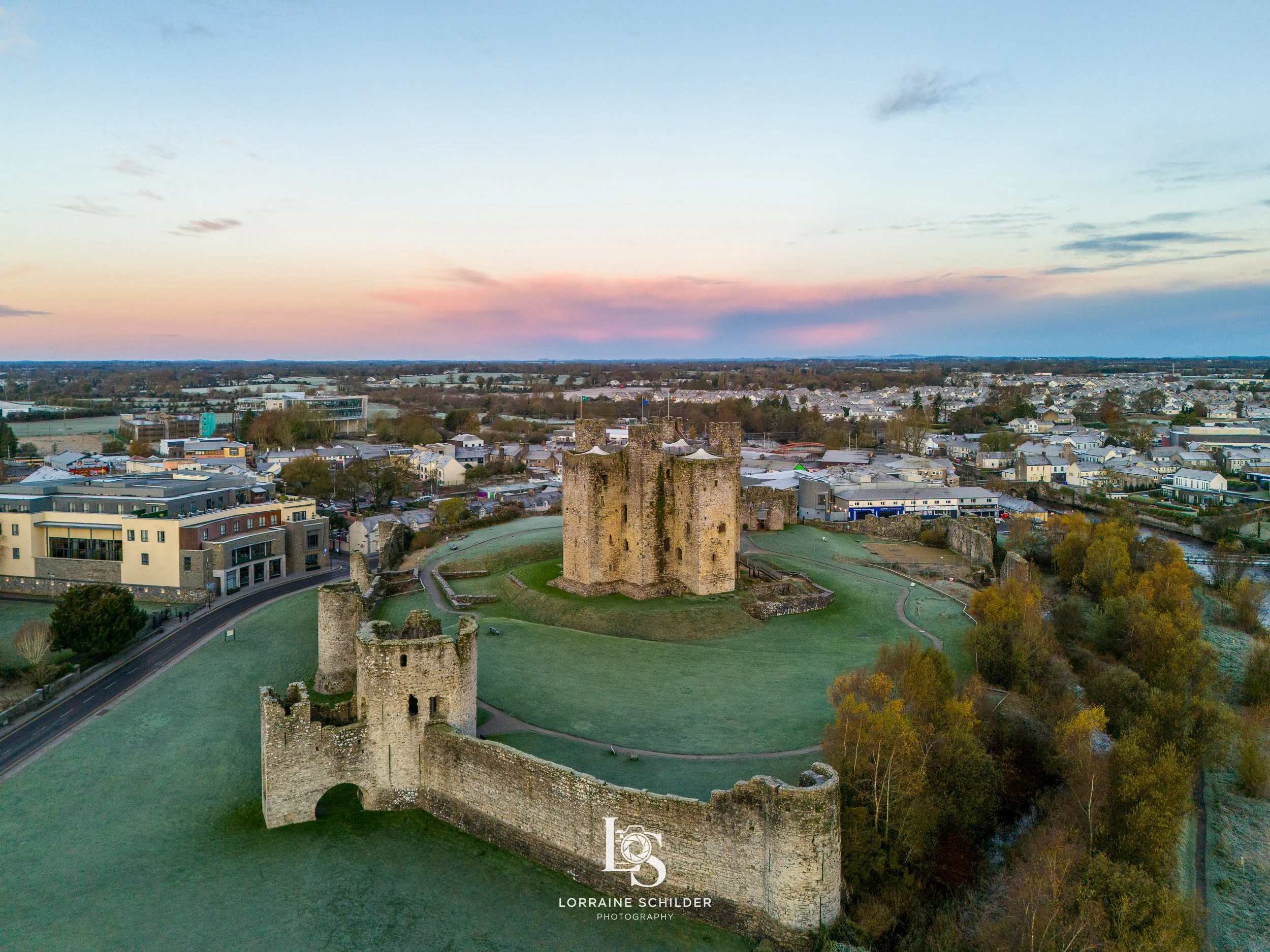 Aerial view of Trim Castle surrounded by green lawns, trees, and a modern Trim town at sunrise.