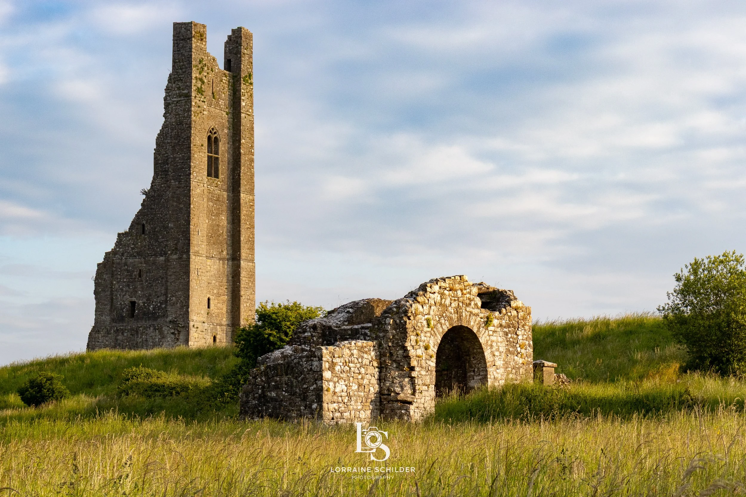 Yellow Steeple & Sheep's Gate with a tall, partially ruined tower and an arched stone entrance, surrounded by grassy fields and a cloudy sky.