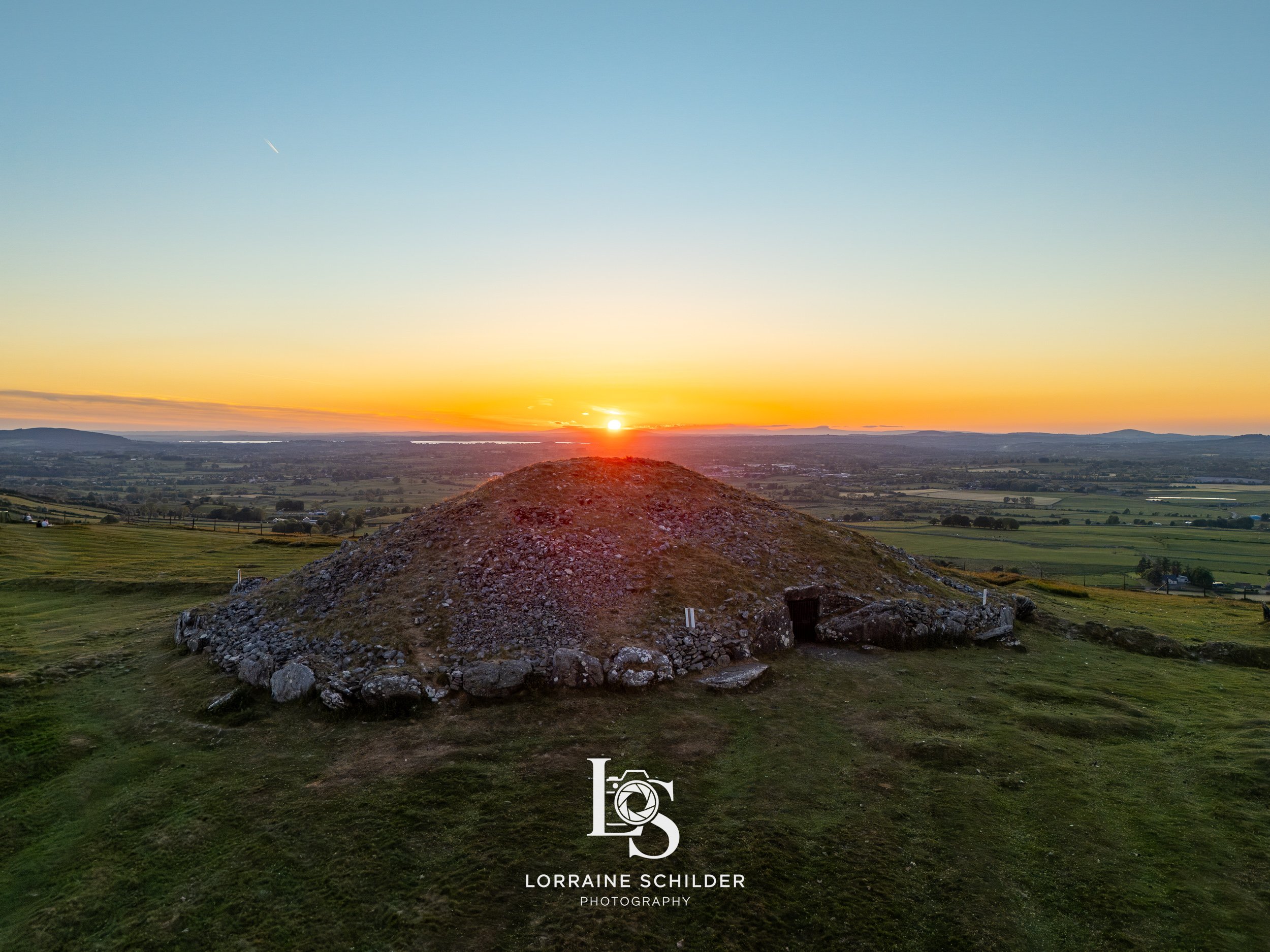 Sunset over a grassy landscape featuring a large, rocky burial mound with a small entrance, with the sun setting near the horizon. Loughcrew, Meath.
