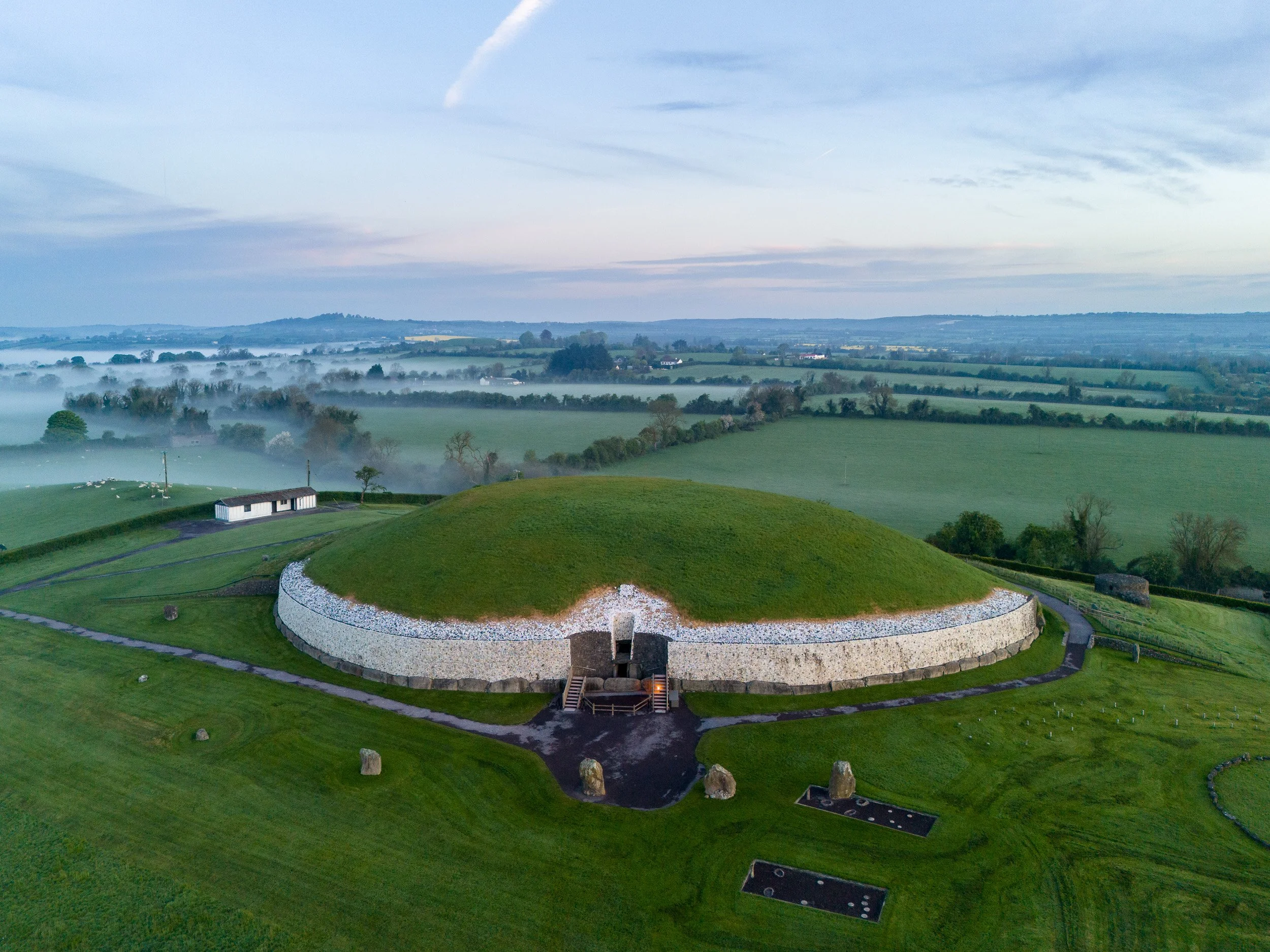 An aerial view of a large historic burial mound surrounded by green fields, with a path leading to its entrance and a small building nearby, under a cloudy sky with a layer of mist in the background.