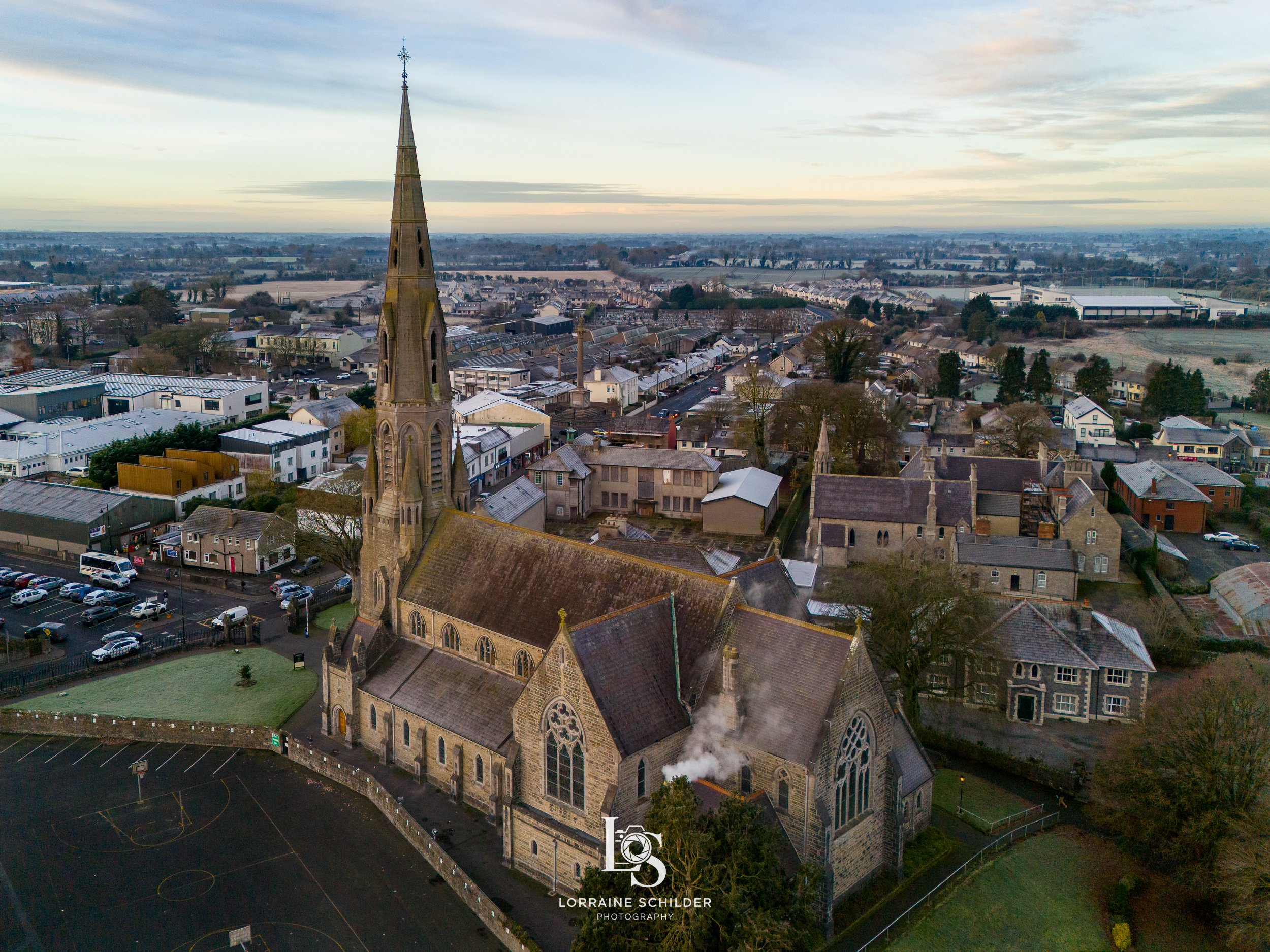 An aerial view St. Patricks church with a tall steeple, surrounded by residential houses, a parking lot, and open fields in the distance during dawn.