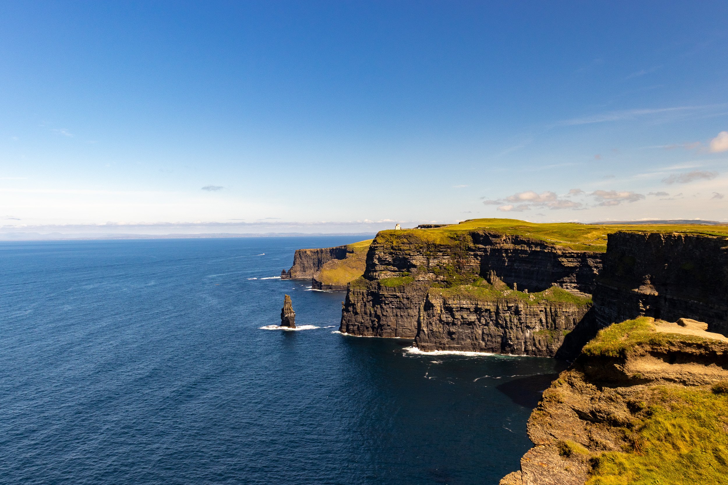 Cliffs along a coastline with green grass on top, dark rocky formations, and a blue ocean extending to the horizon under a clear sky. Cliffs of Moher, Clare.