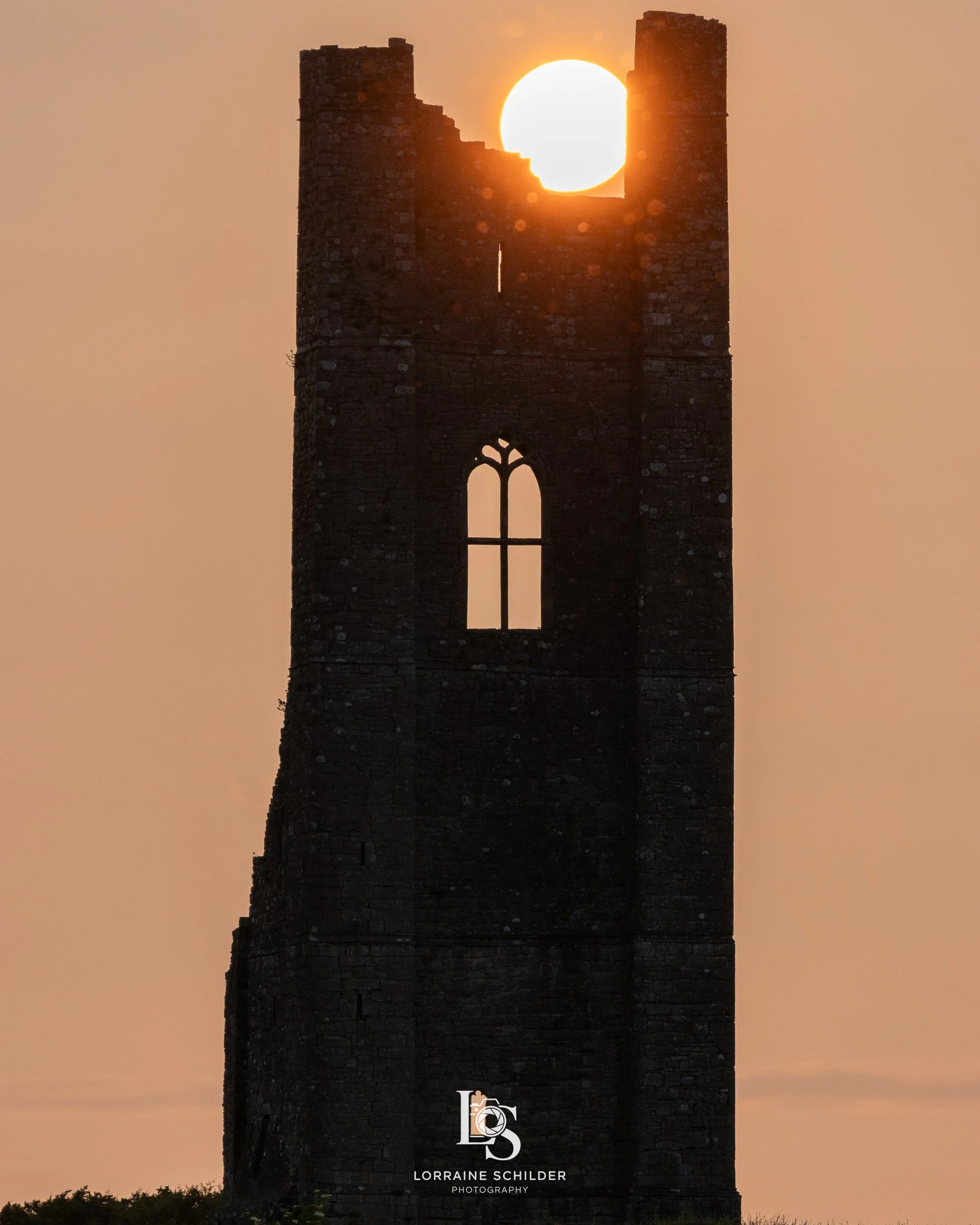 Sun setting behind a tall, weathered stone tower with arched windows, creating a silhouette against the orange sky.
