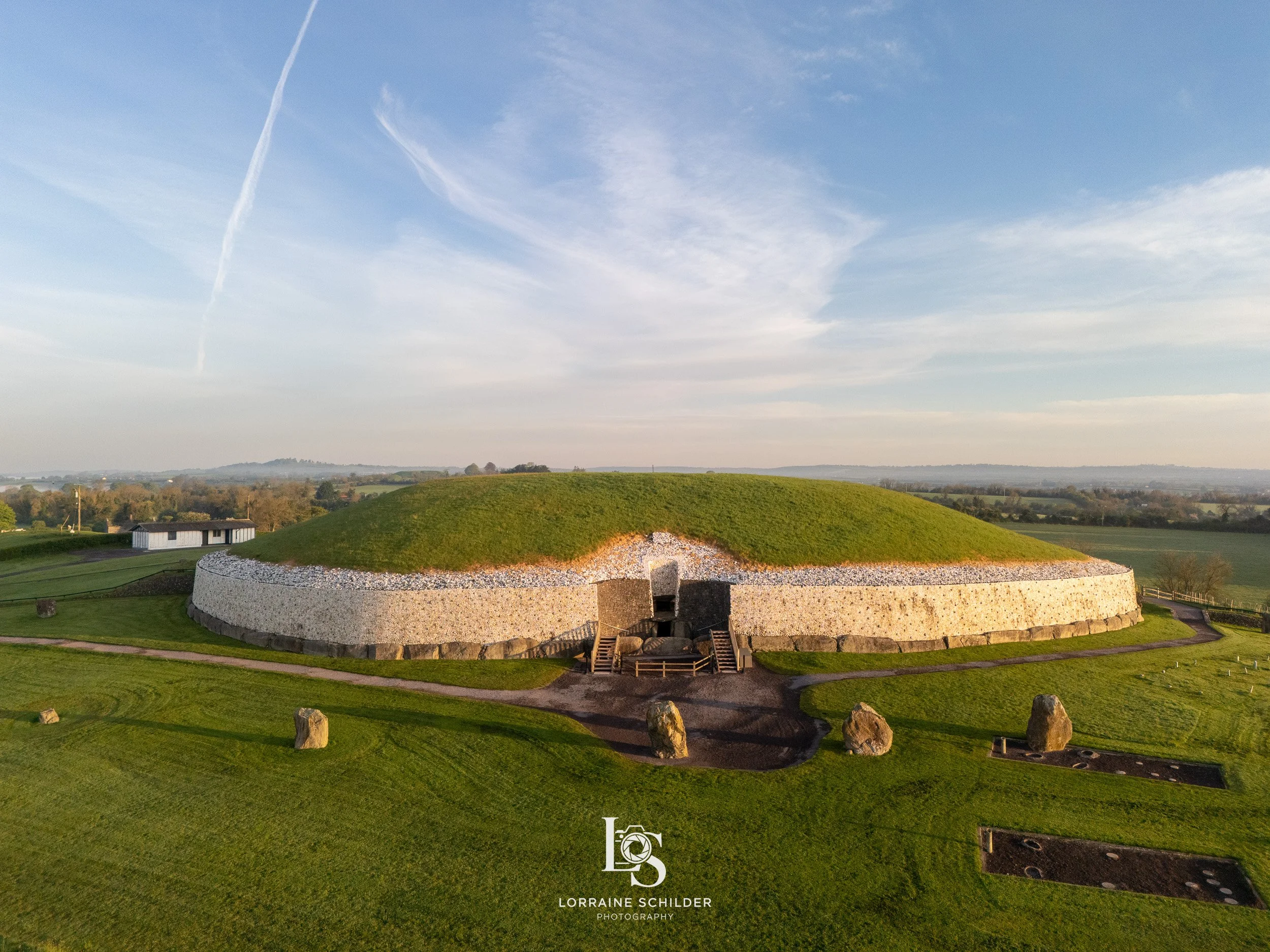 A large ancient burial mound or tumulus with grass covering the top and stone walls around the sides, set in a green landscape with a clear sky overhead.  Newgrange, Meath.