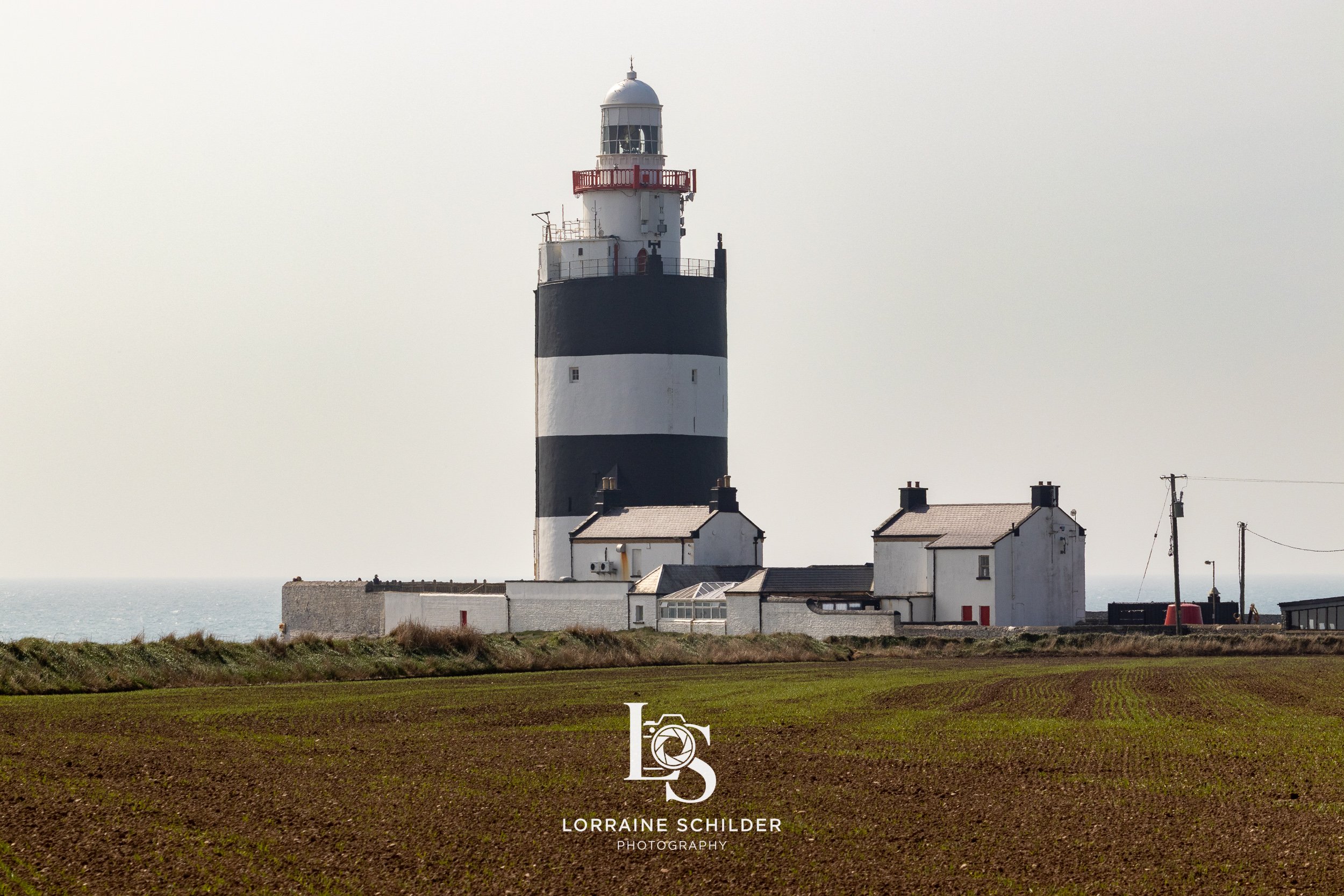 A black and white striped lighthouse with a small white building complex at its base on a grassy land near the coast, overcast sky. Wexford.