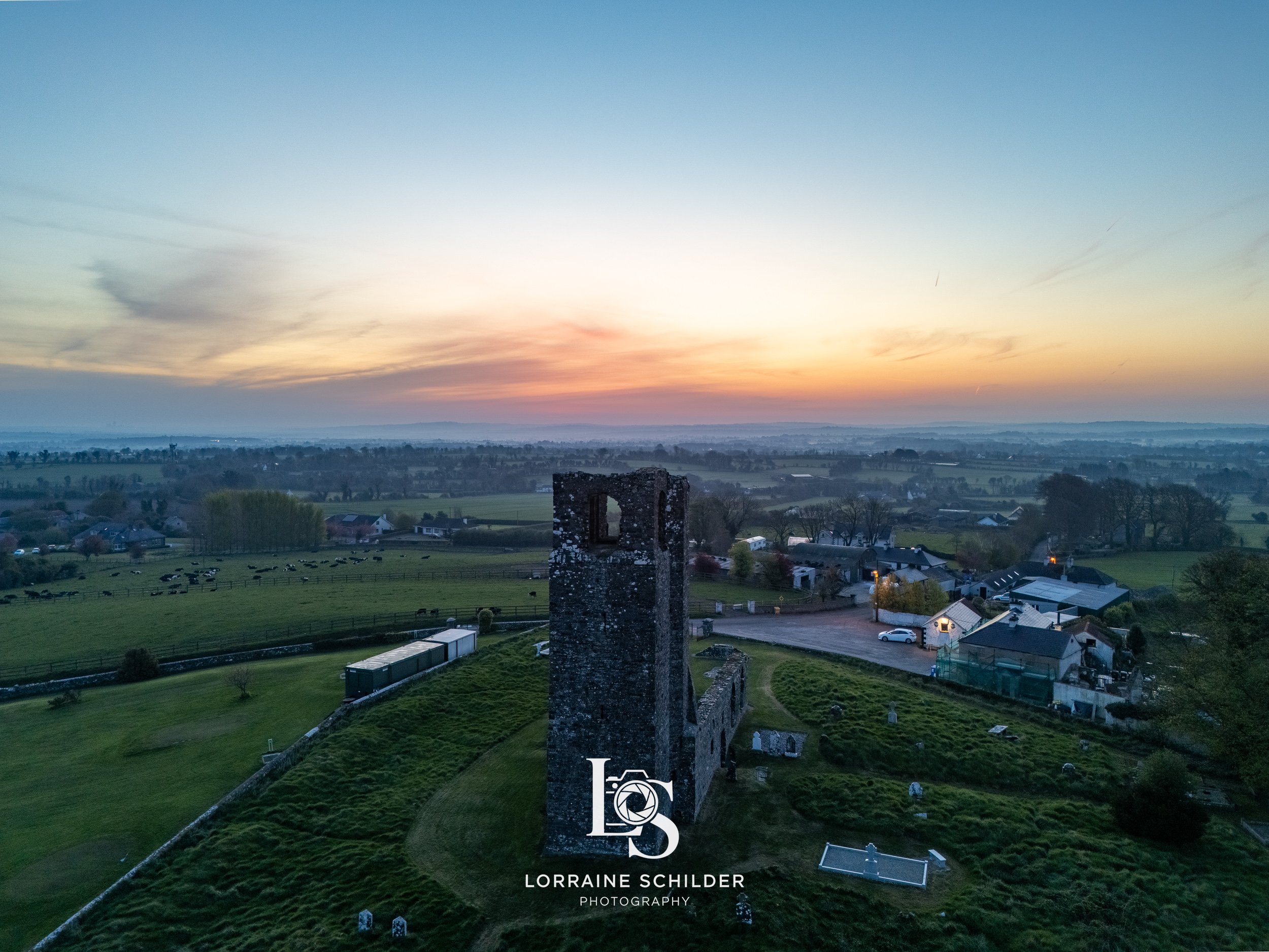 An aerial view of a rural landscape during sunset, featuring an old stone tower in the foreground, green fields with grazing cows, and a small village with houses and farm buildings. Skyrne, Meath.