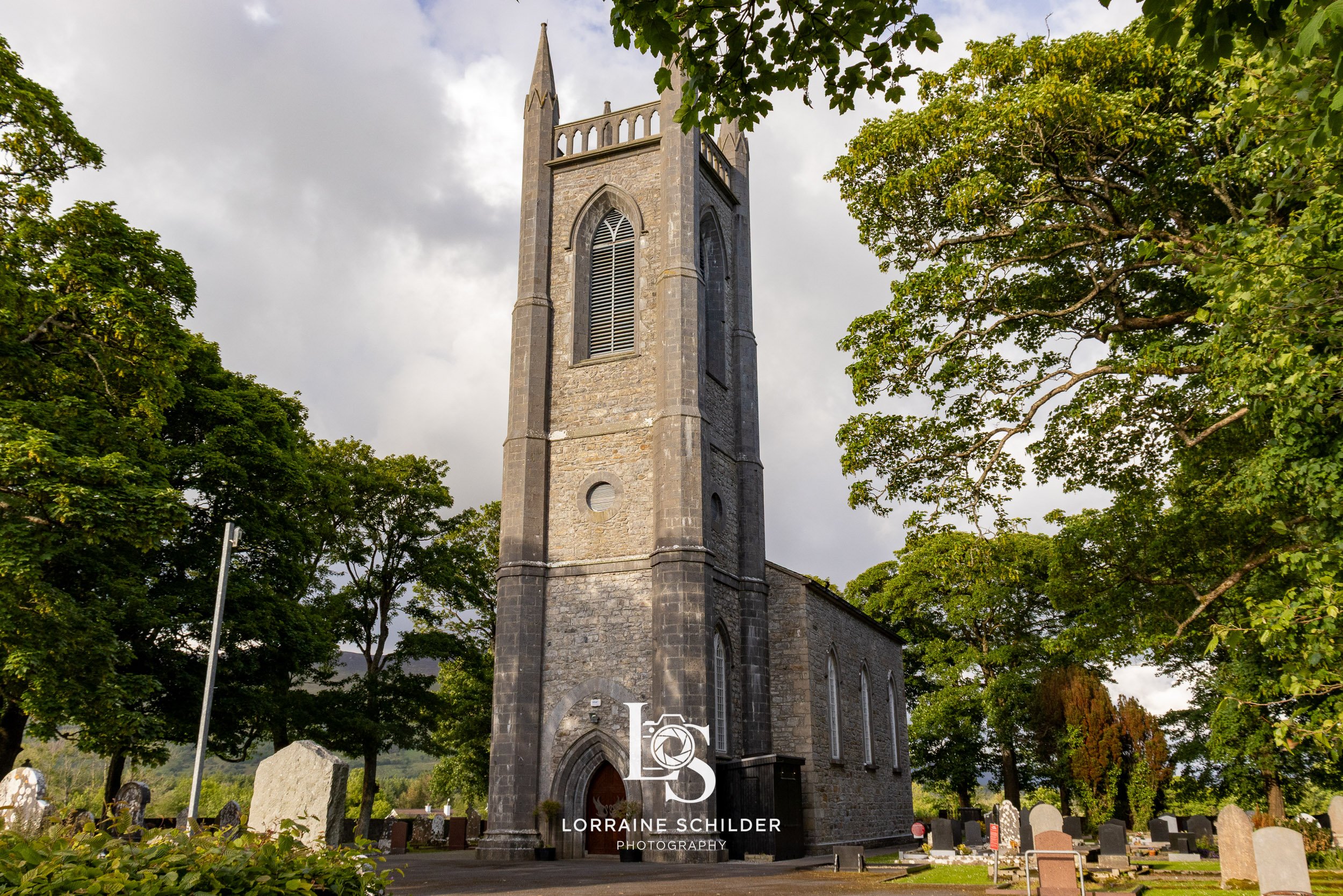 A stone church with a tall, pointed steeple surrounded by green trees and a graveyard with tombstones. Sligo.