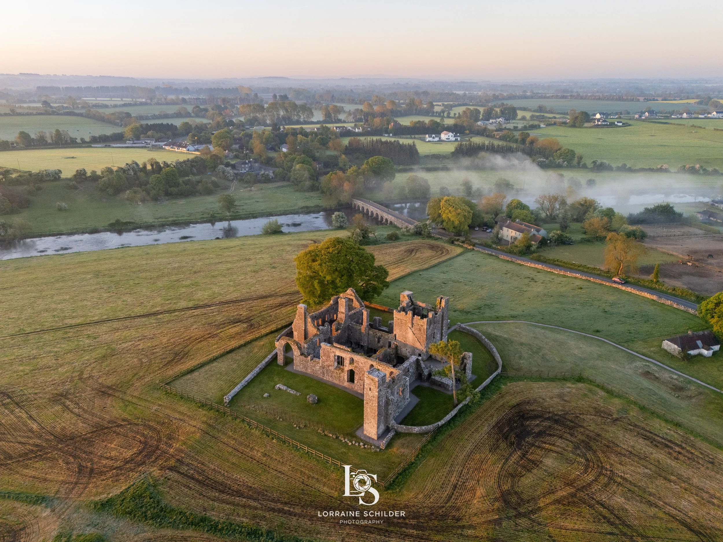 Aerial view of an ancient stone castle ruins surrounded by green fields, trees, a river, and bridge in the background, during sunrise. Bective Abbey, Meath.