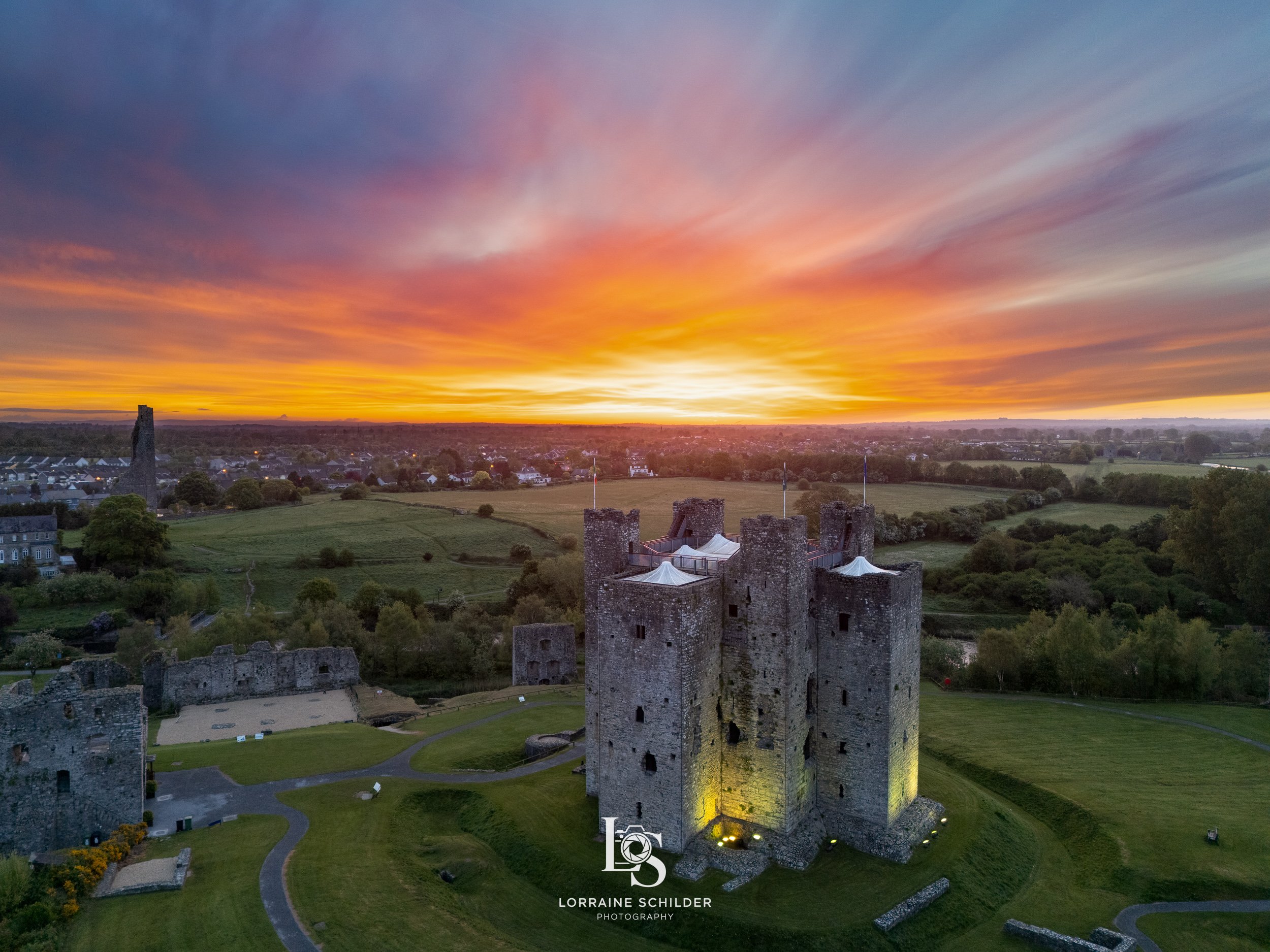 Aerial view of Trim castle at sunrise with illuminated stone walls, surrounded by green parks, and fields, under a colorful sky with orange, yellow, purple, and blue clouds.