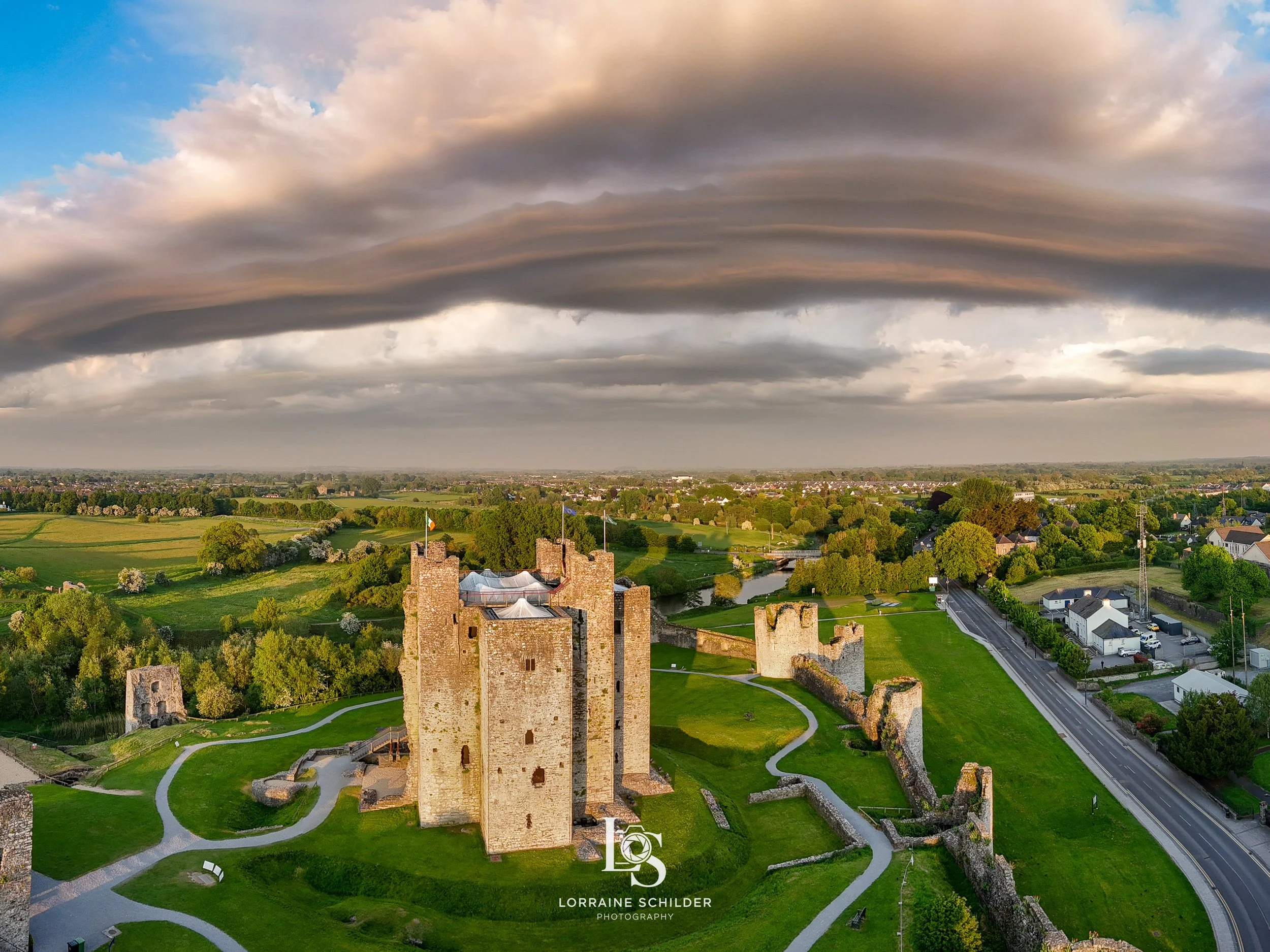An aerial view of a Trim Castle surrounded by green fields, trees, a river, and a nearby town of Trim  under a dramatic cloudy sky at sunset.