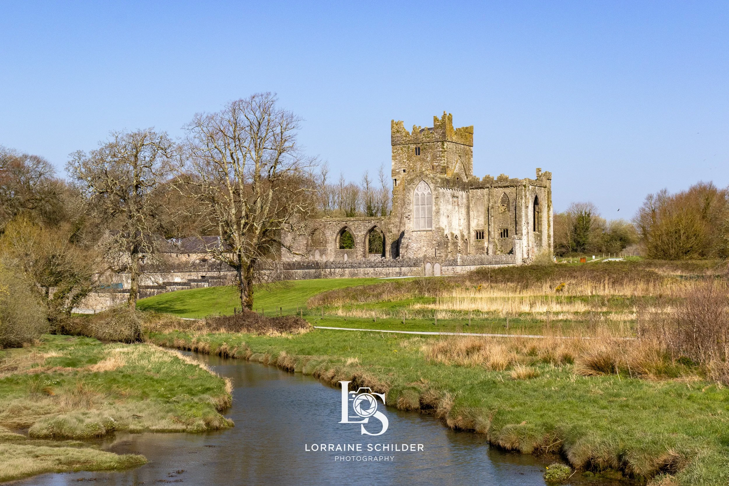 A historic stone castle with a tall central tower and arched windows, situated on a grassy landscape with leafless trees and a small river running in the foreground, under a clear blue sky. Wexford.