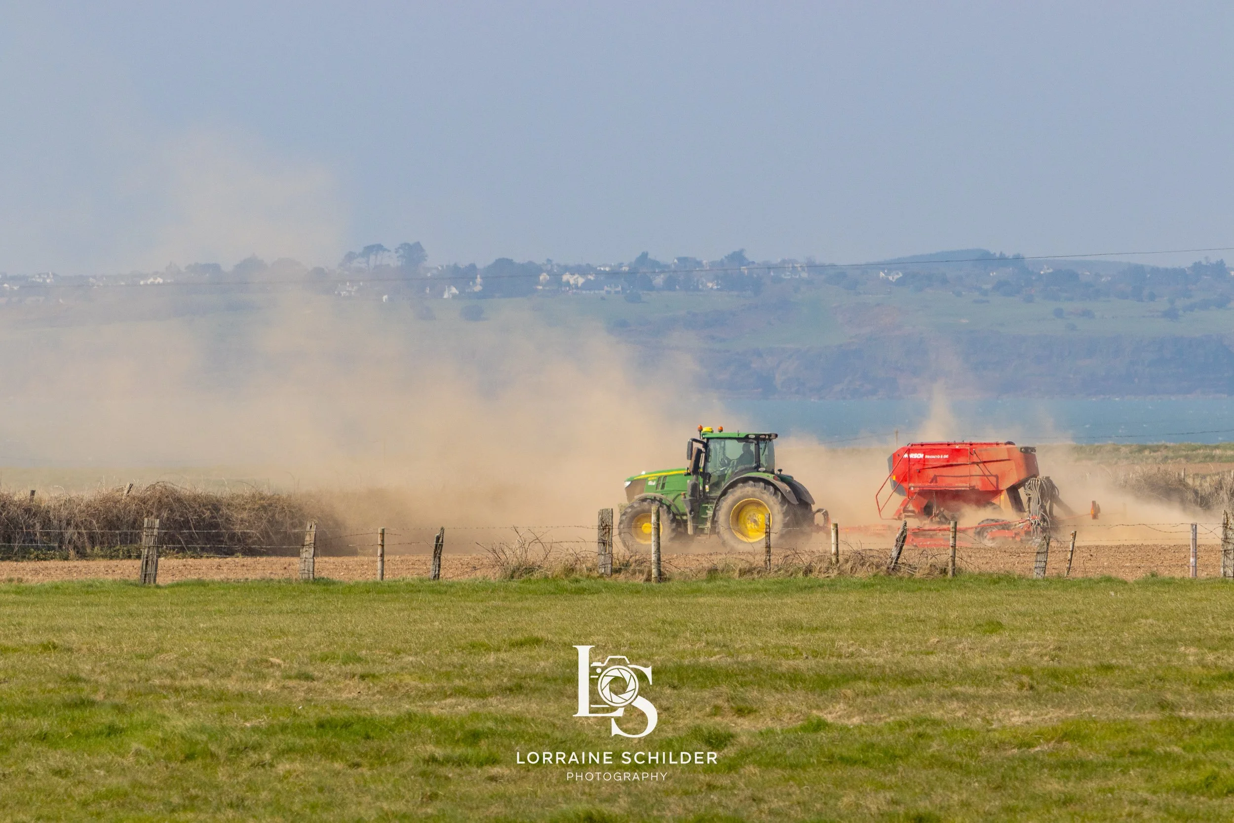 A green tractor with a red attached farming implement working in a field, kicking up dust in a rural landscape with grass, a fence, and distant hills. Wexford.